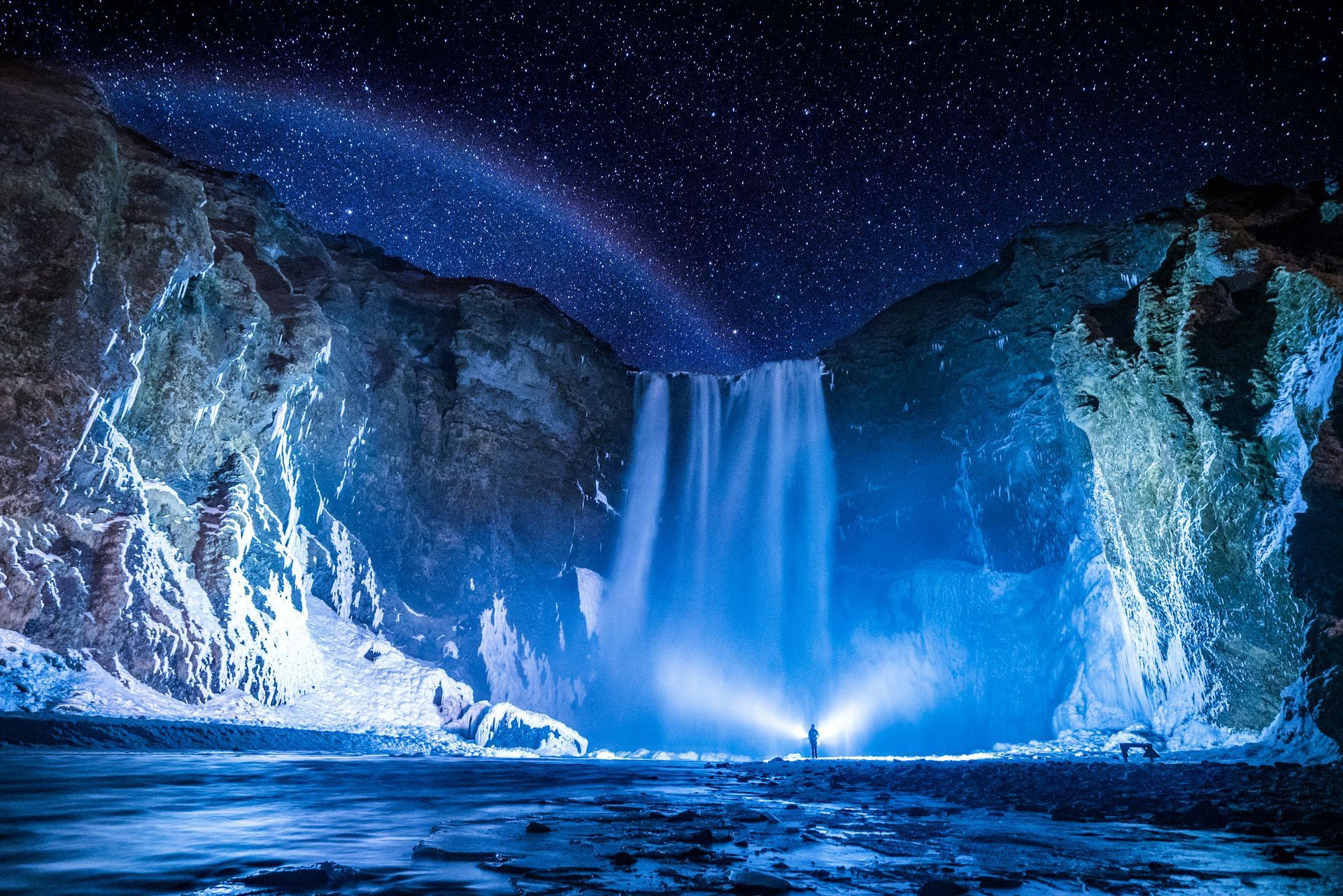 Skógafoss Waterfall under starry night sky, illuminated with blue light. Figures stand in front of the falls in Iceland.