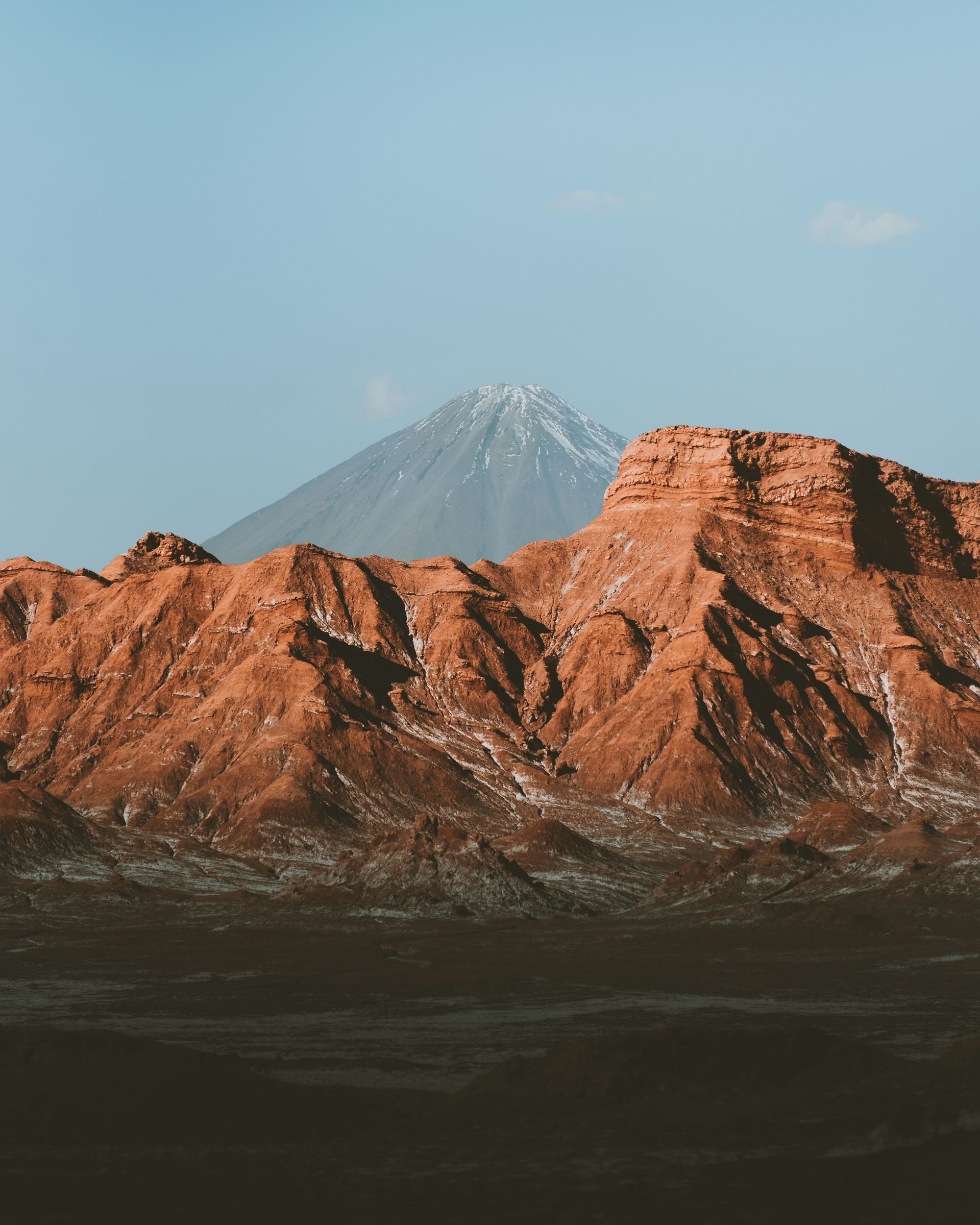 Brown rock formations with a snowy mountain peak and a blue sky in the Atacama Desert, Chile.
