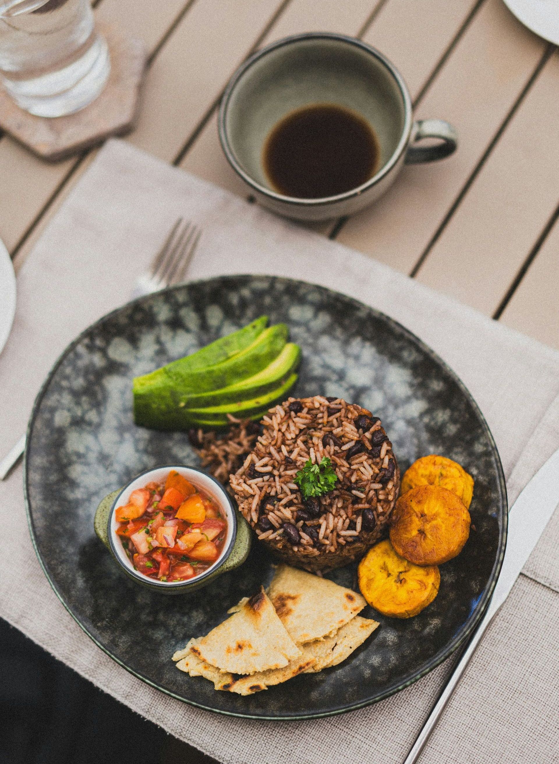 A plate of Costa Rican Gallo Pinto with avocado, plantains, salsa, and tortillas, next to a coffee cup.