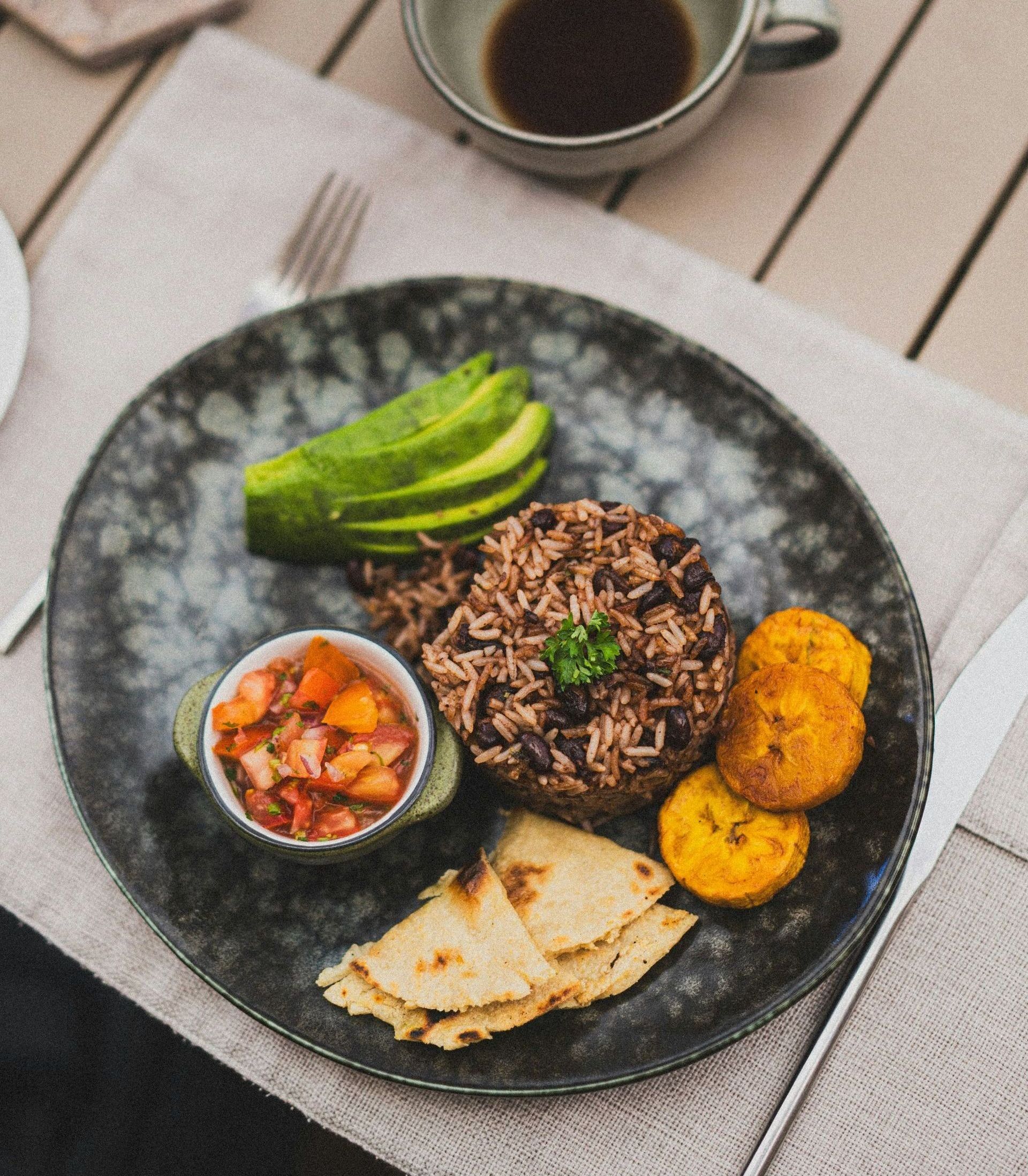 A plate of Costa Rican Gallo Pinto with avocado, plantains, salsa, and tortillas, next to a coffee cup.