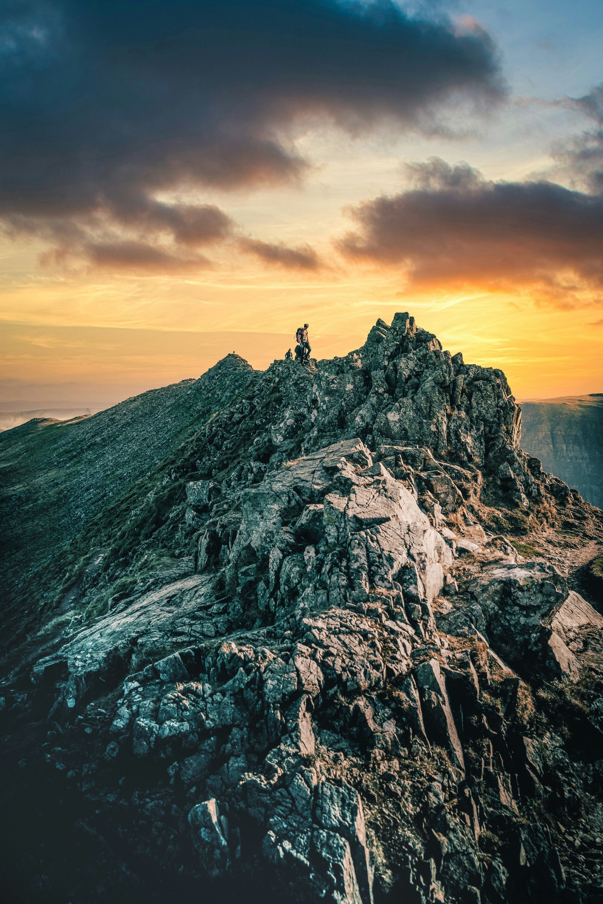 Person atop a rocky mountain peak at sunset. Orange and yellow sky; rugged terrain in the Lake District, England.