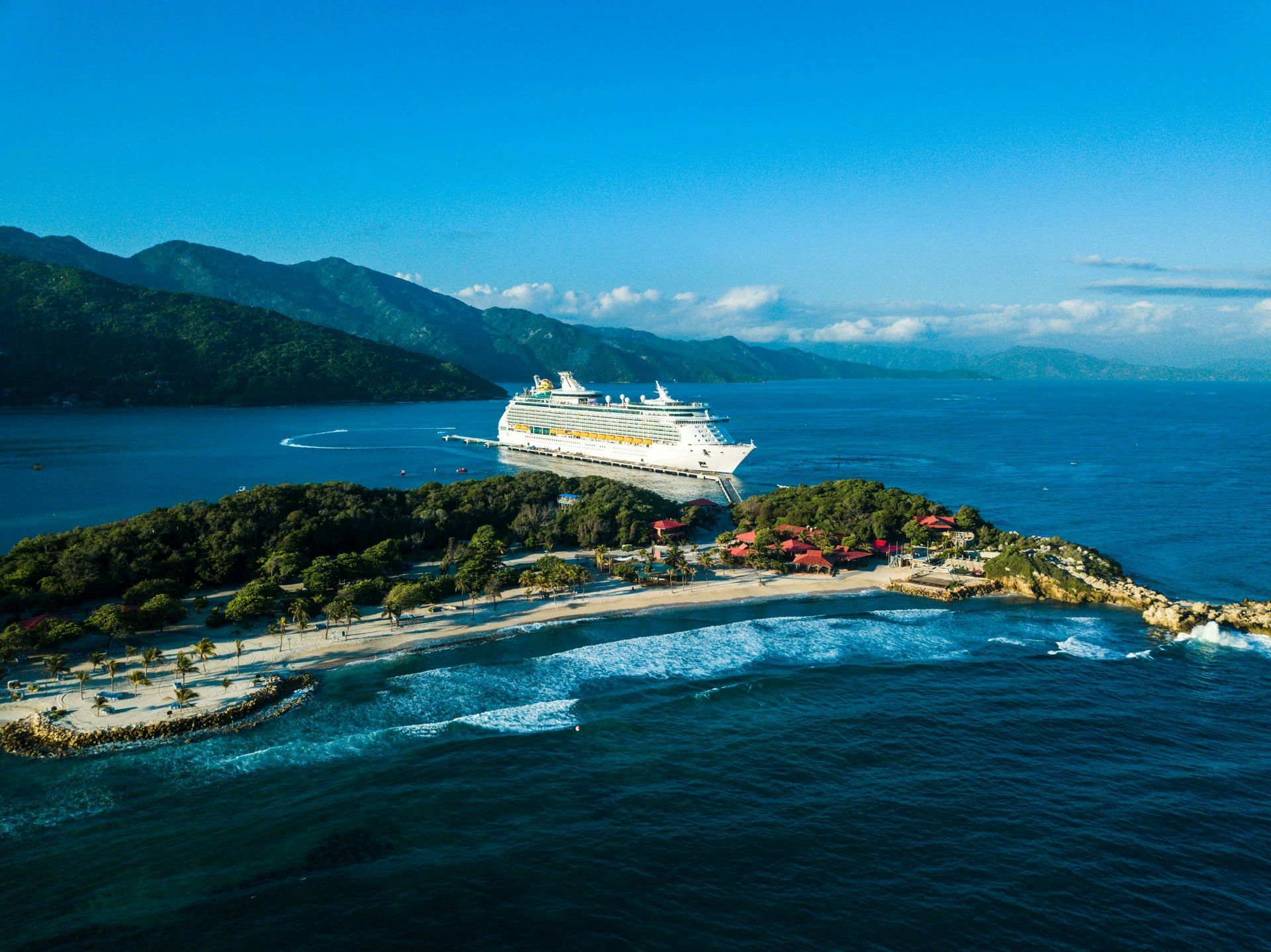 Cruise ship docked at a tropical island with white sand beach, green trees, and blue water.