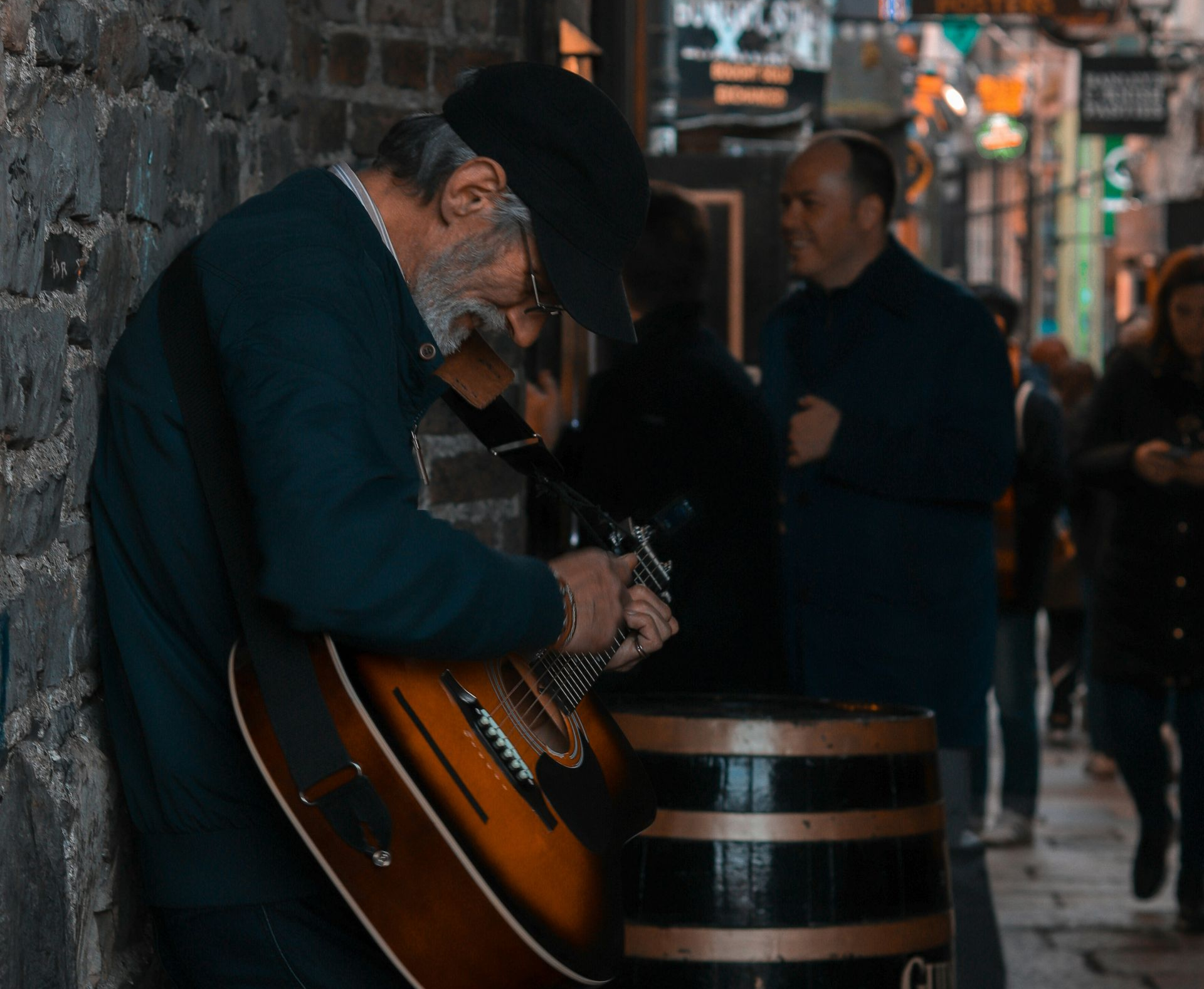 Man playing guitar in an alley, leaning against a brick wall, with passersby in the background in Dublin, Ireland.
