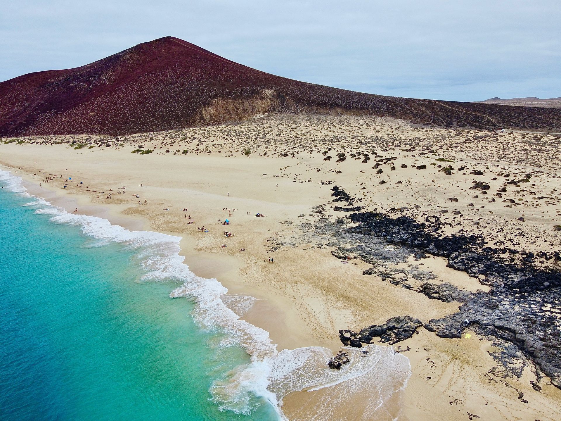 Beach with turquoise water, sand, and dark volcanic rock, backed by a dark red volcanic hill on the Canary Islands, Spain.
