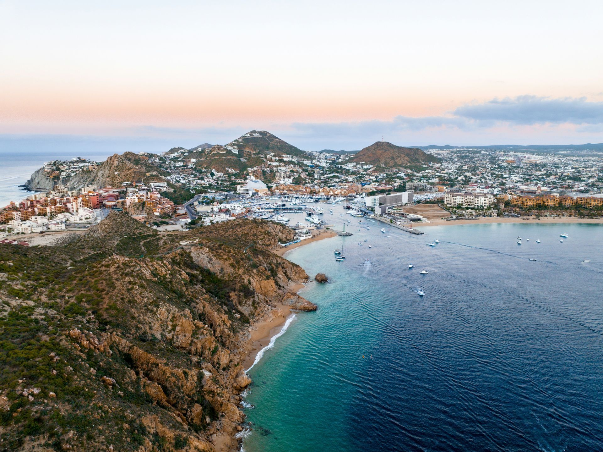 Aerial view of Cabo San Lucas coastline with buildings and marina, showing the ocean and beach meeting the rocky shore.