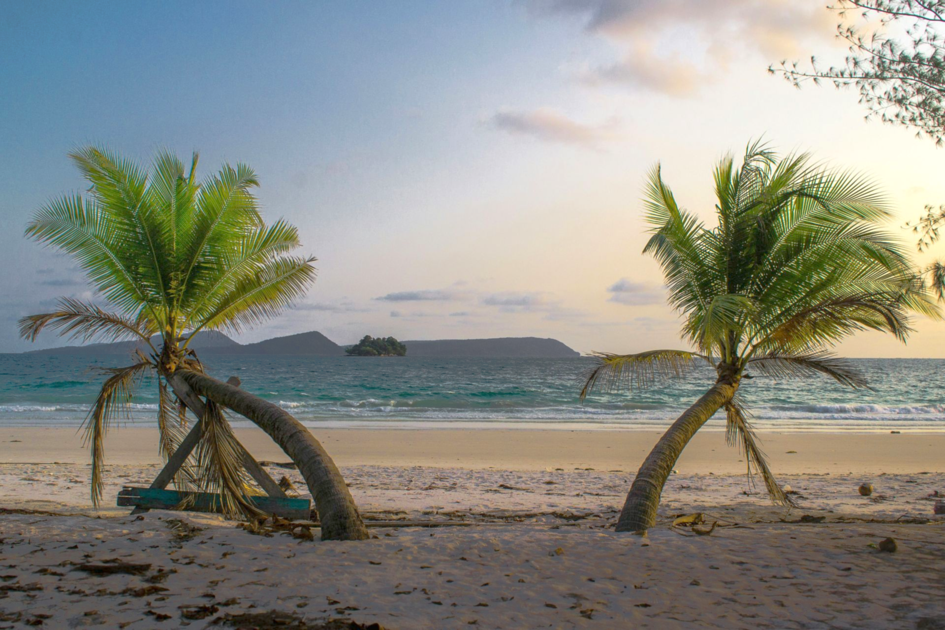 Two palm trees on a sandy beach with a calm sea and island in the distance in Koh Rong, Cambodia.
