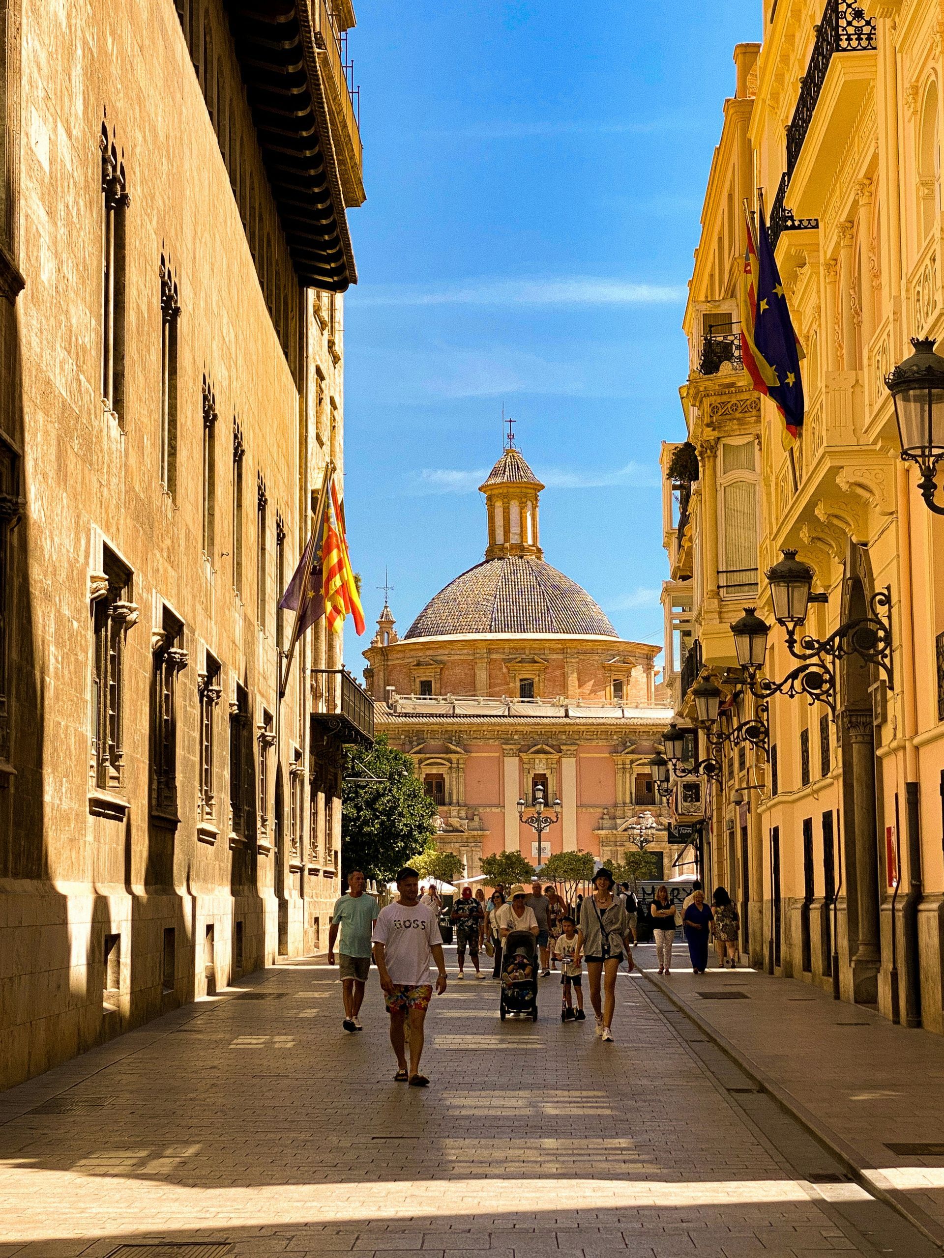Narrow street in Valencia, Spain, with pedestrians walking towards a pink domed building under a blue sky.