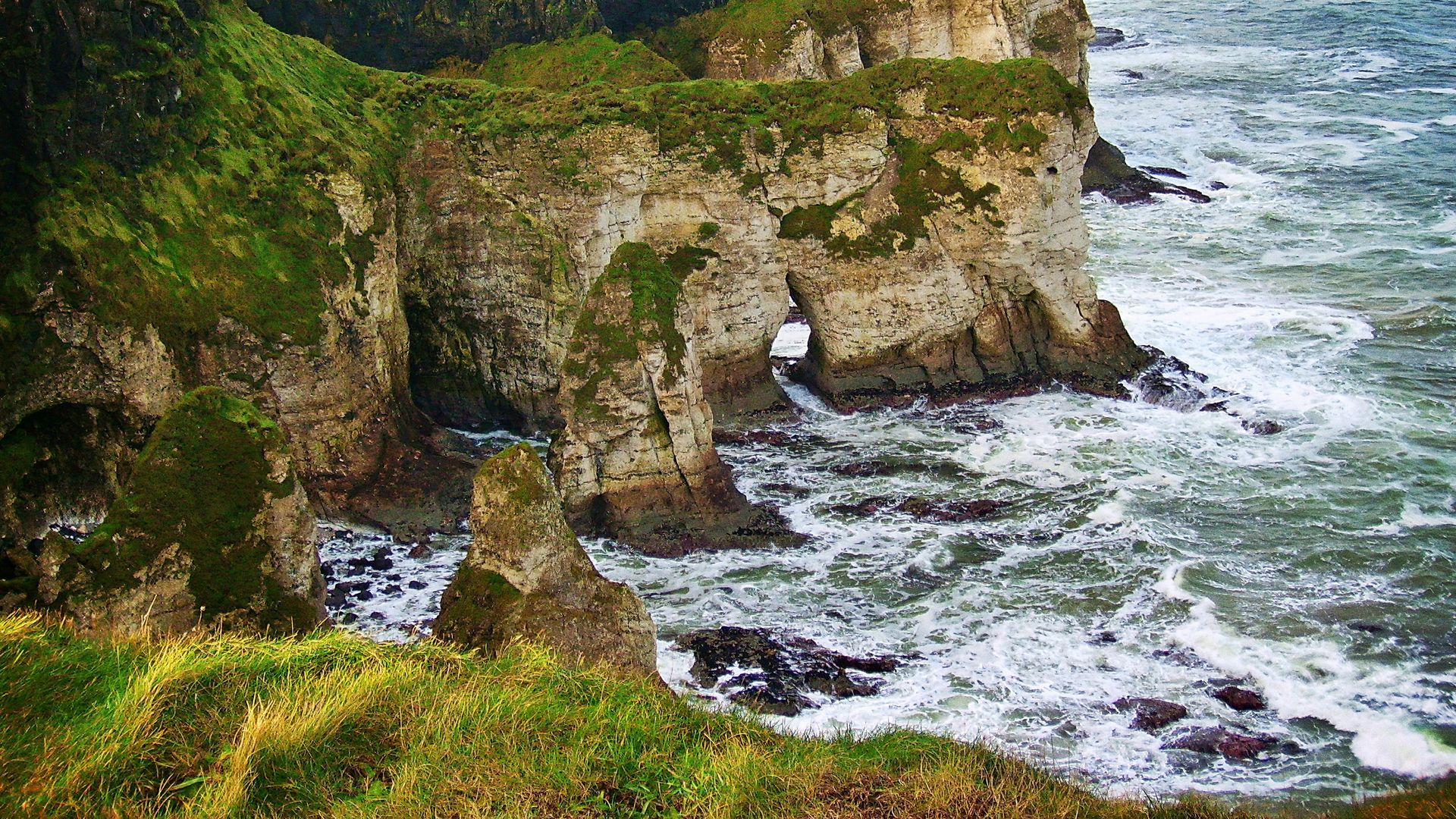 Rocky cliffs covered in green vegetation meet turbulent ocean waves in Northern Ireland.