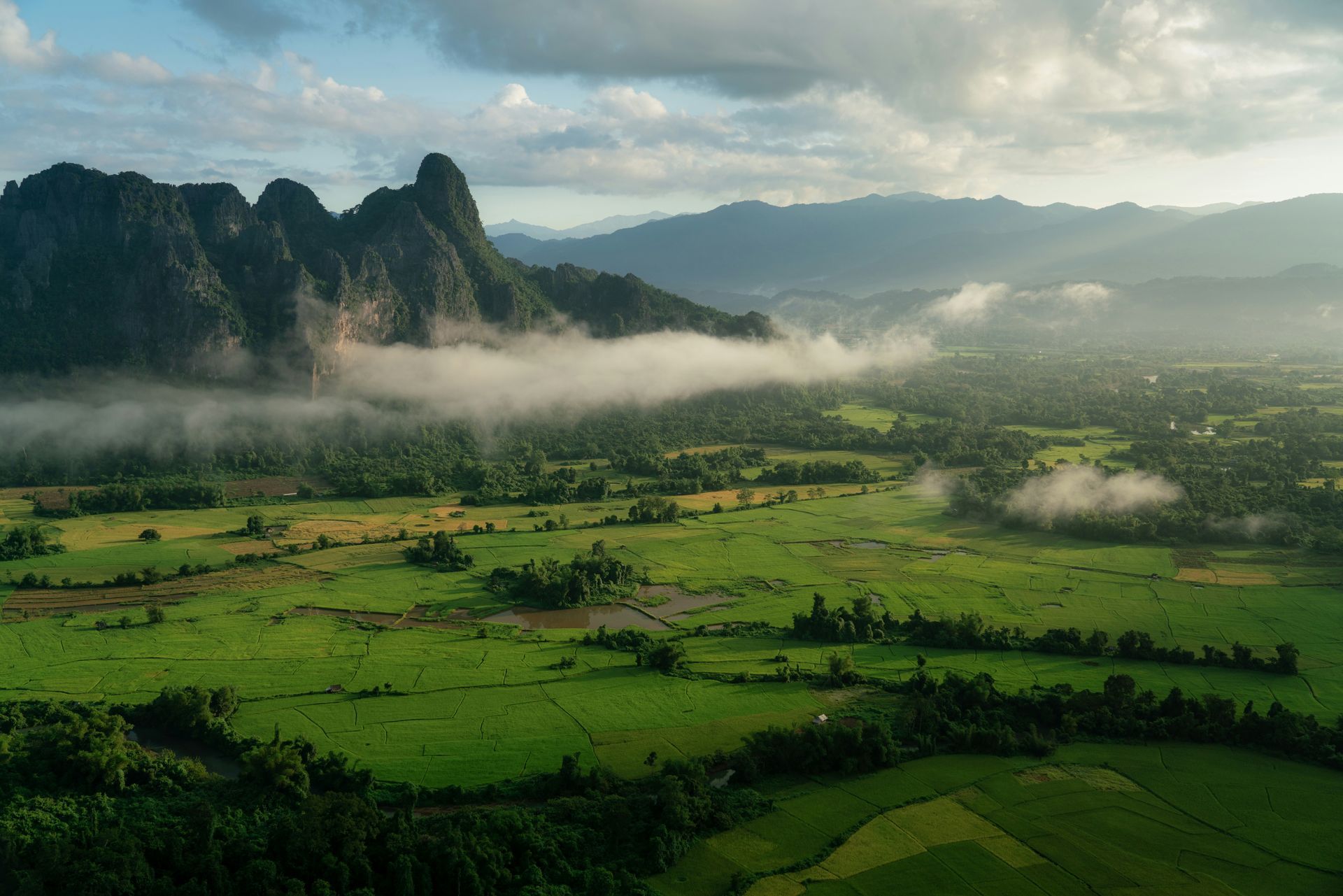 Green valley with low-lying clouds, tall limestone peaks in the background in Vang Vieng, Laos.