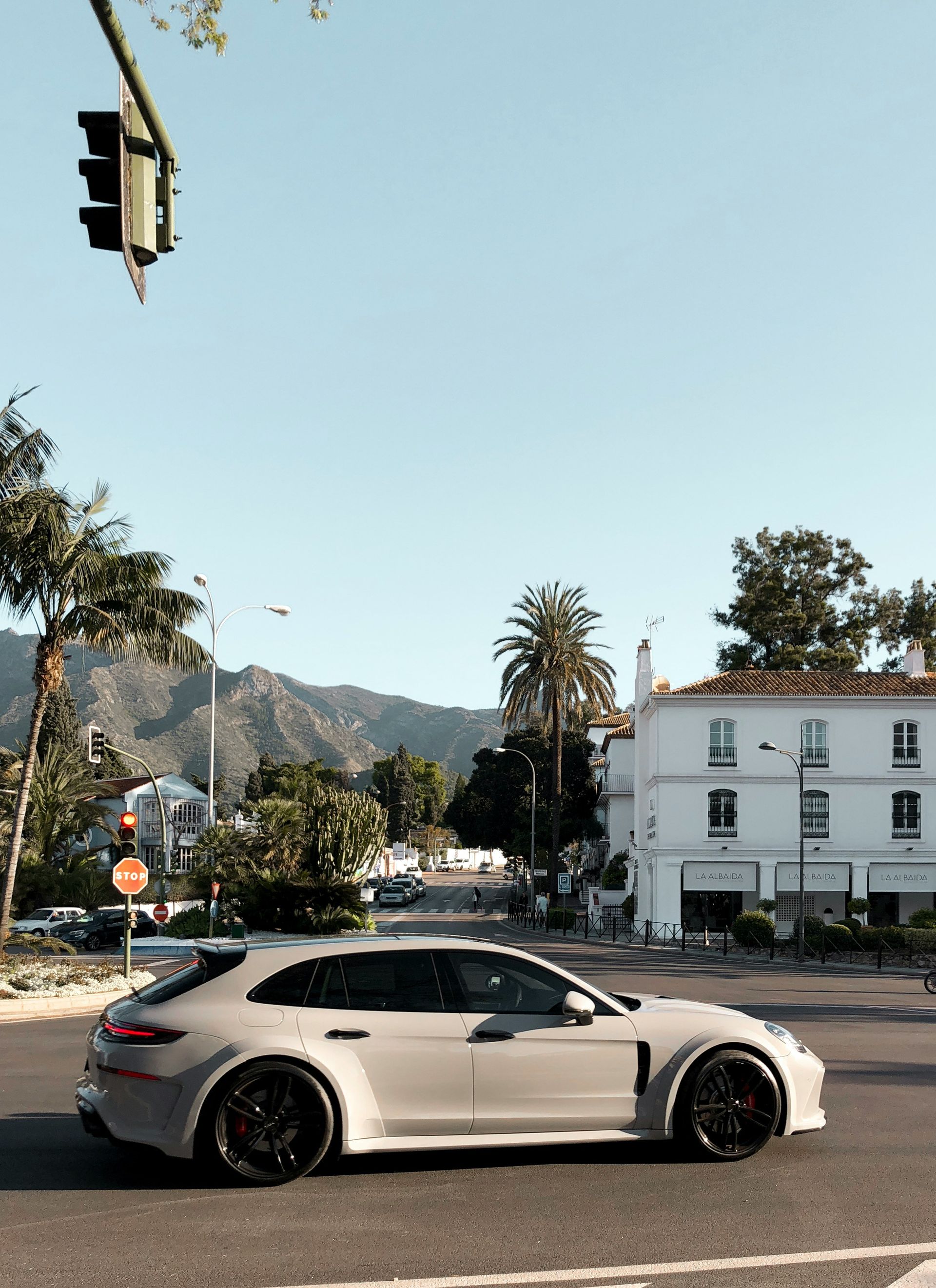 Gray Porsche Panamera driving on a street with buildings and palm trees under a blue sky in Marbella, Spain.