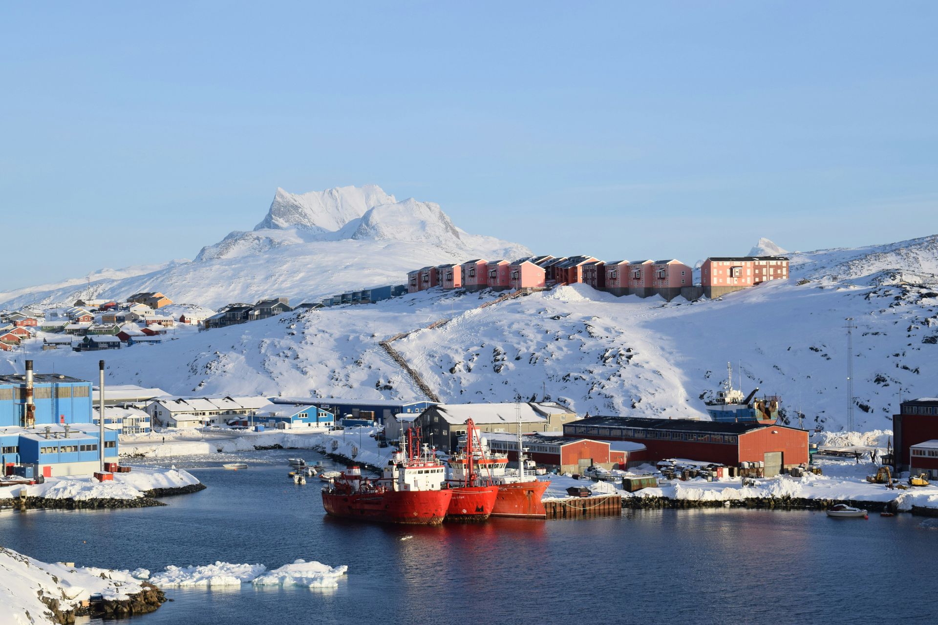 Snowy harbor scene in Nuuk, Greenland: red ships, buildings, and snow-covered mountain under a clear blue sky.