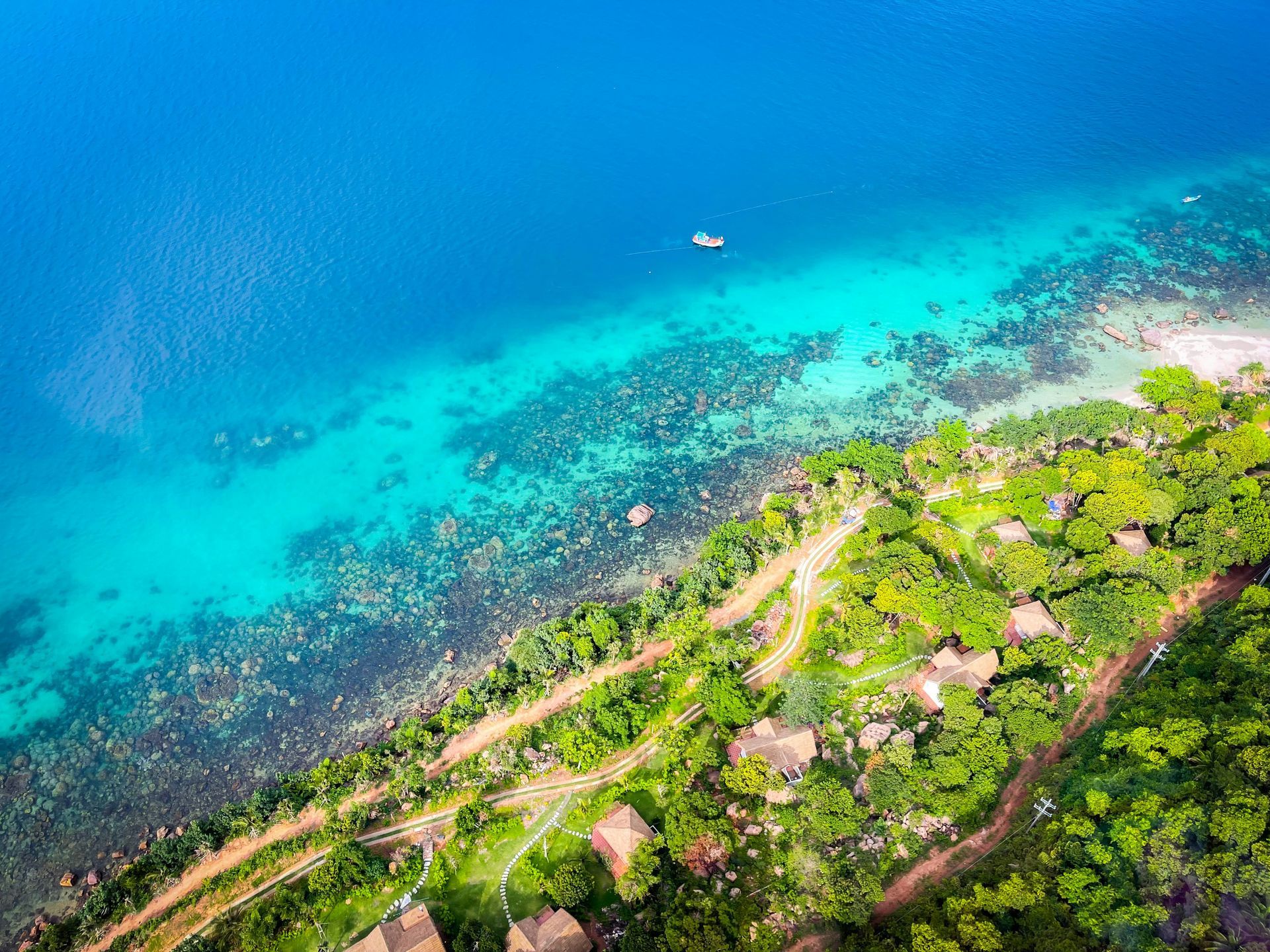 Aerial view of a tropical coastline with vibrant blue water, a beach, and lush green trees in Phú Quốc, Vietnam.