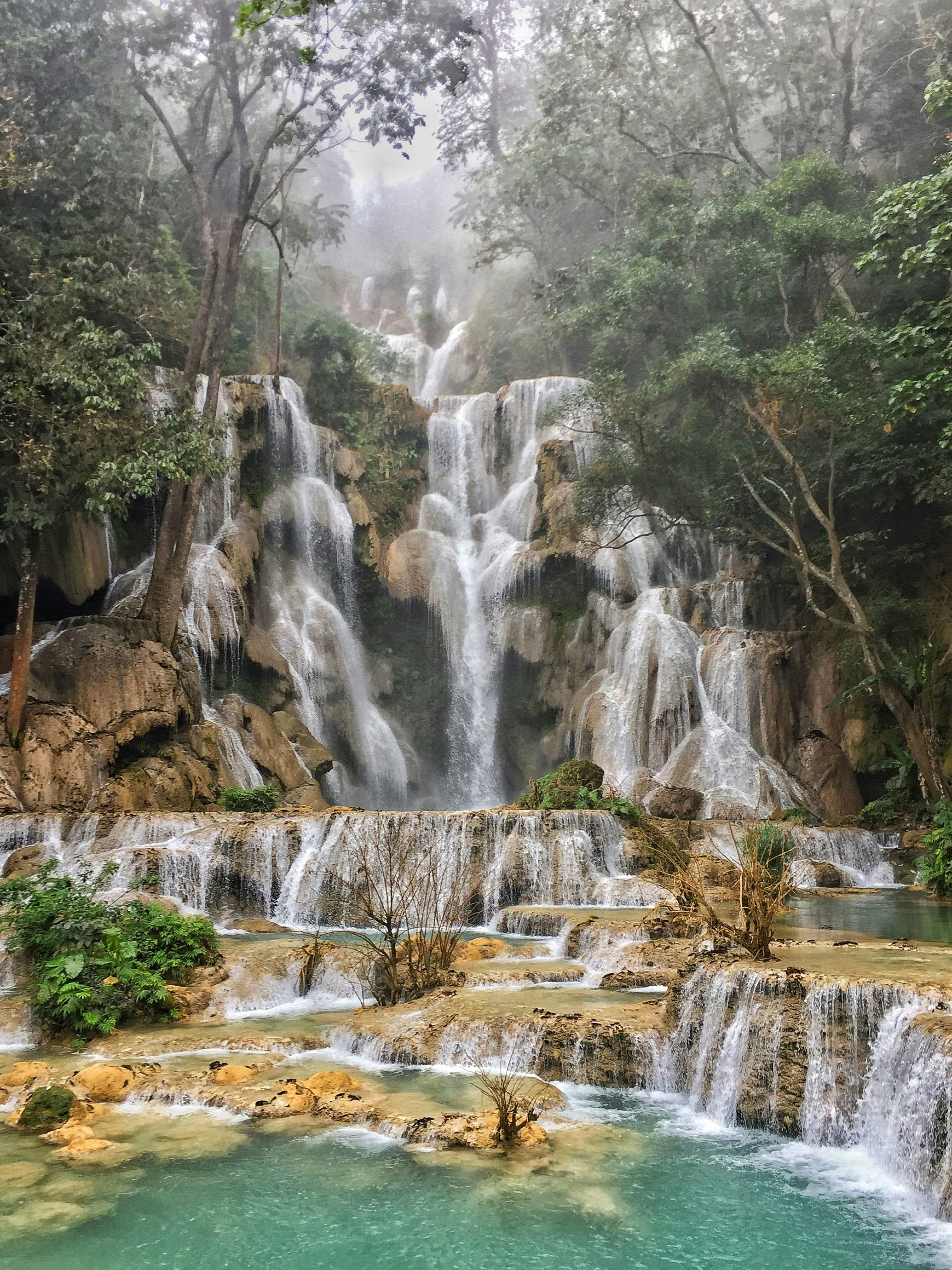 Kuang Si Falls cascading down layered rocks into a turquoise pool, surrounded by green forest in Luang Prabang, Laos.
