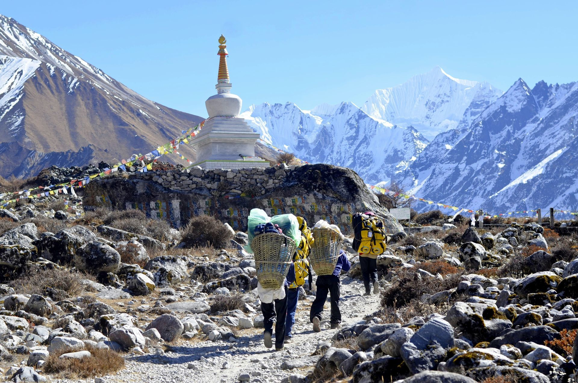 People hiking on a path, carrying large packs, near a white stupa in front of the Himalayas in Langtang, Nepal.