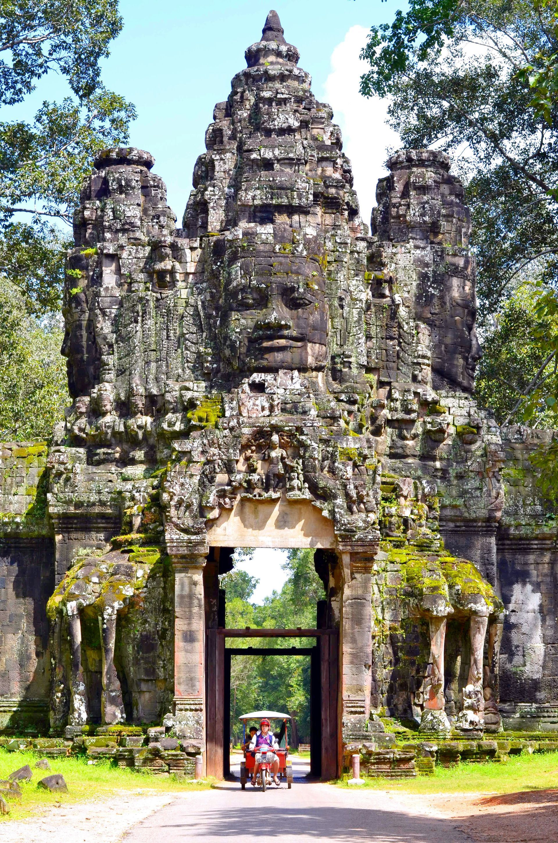 Stone gate with carved faces at Angkor Thom, Cambodia. A tuk-tuk drives through.