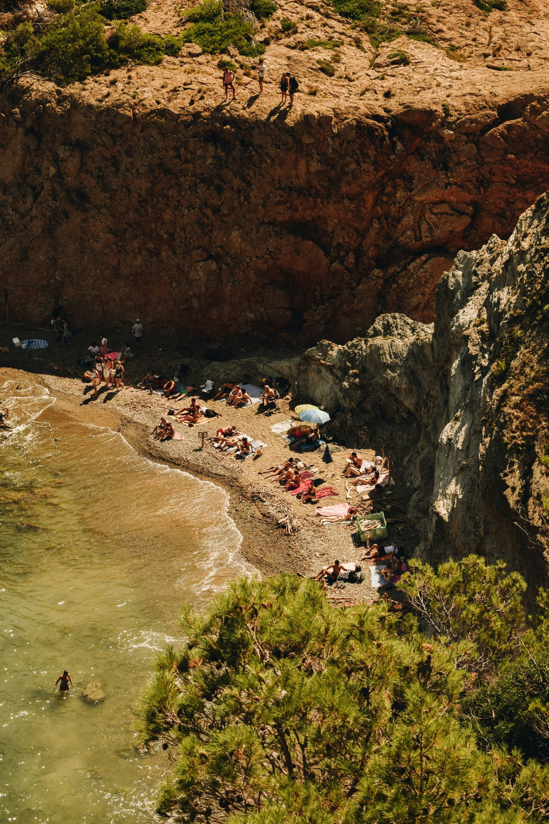 Beach with people sunbathing, rocky cliffs, and turquoise water on Costa Brava, Spain.