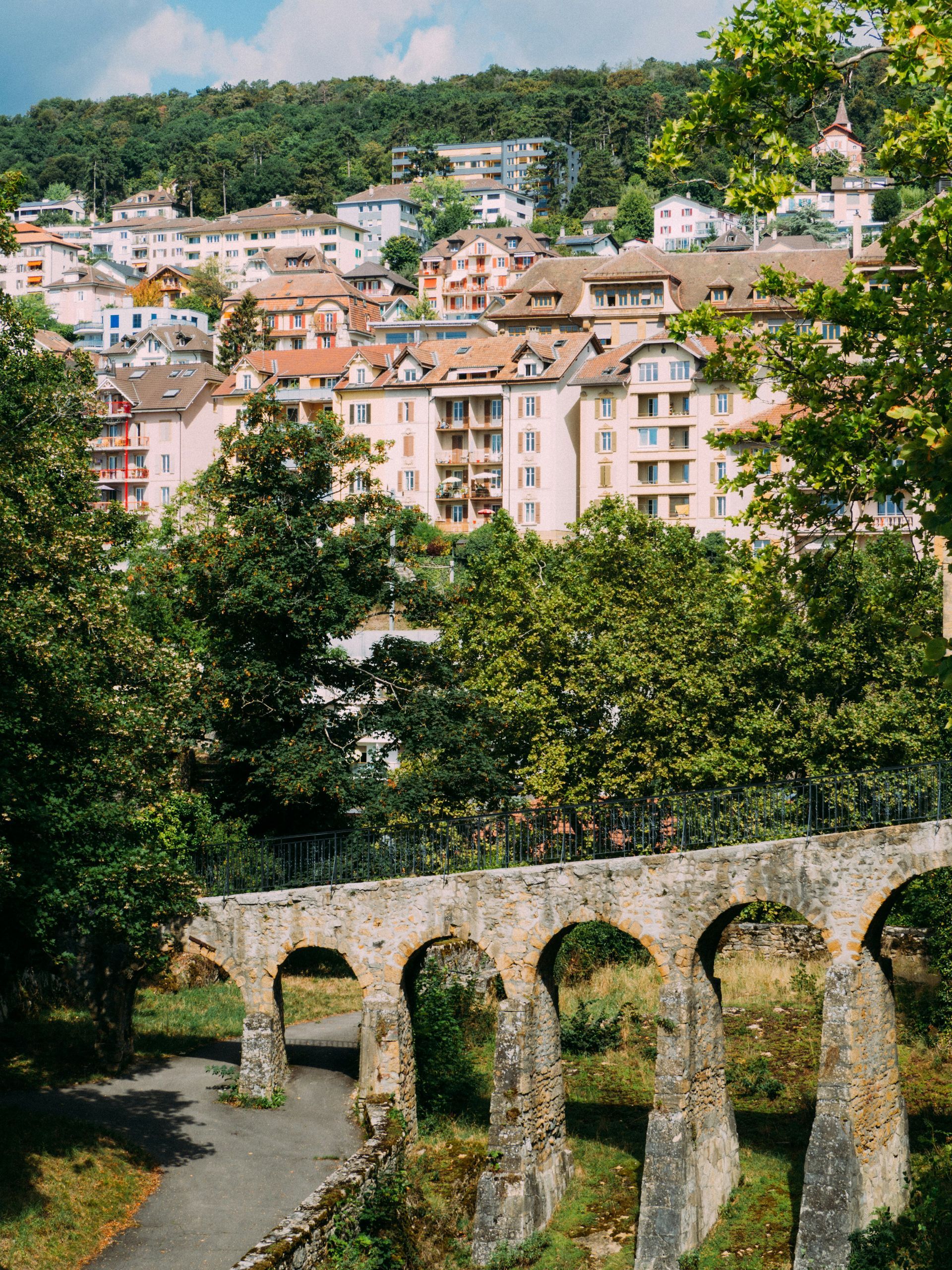 Stone arched bridge in foreground, town on a hillside in the background, trees throughout in Neuchâtel, Switzerland.