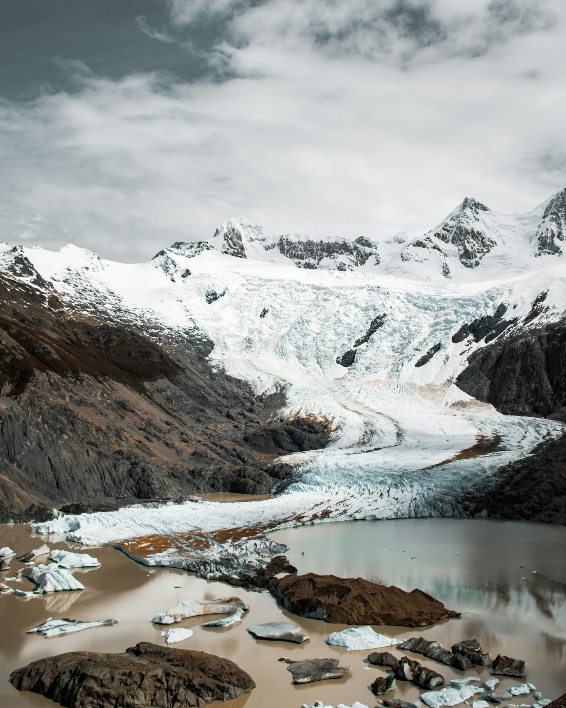 Glacier against snowy mountain, brown lake with ice chunks, cloudy sky in El Chaltén, Argentina.