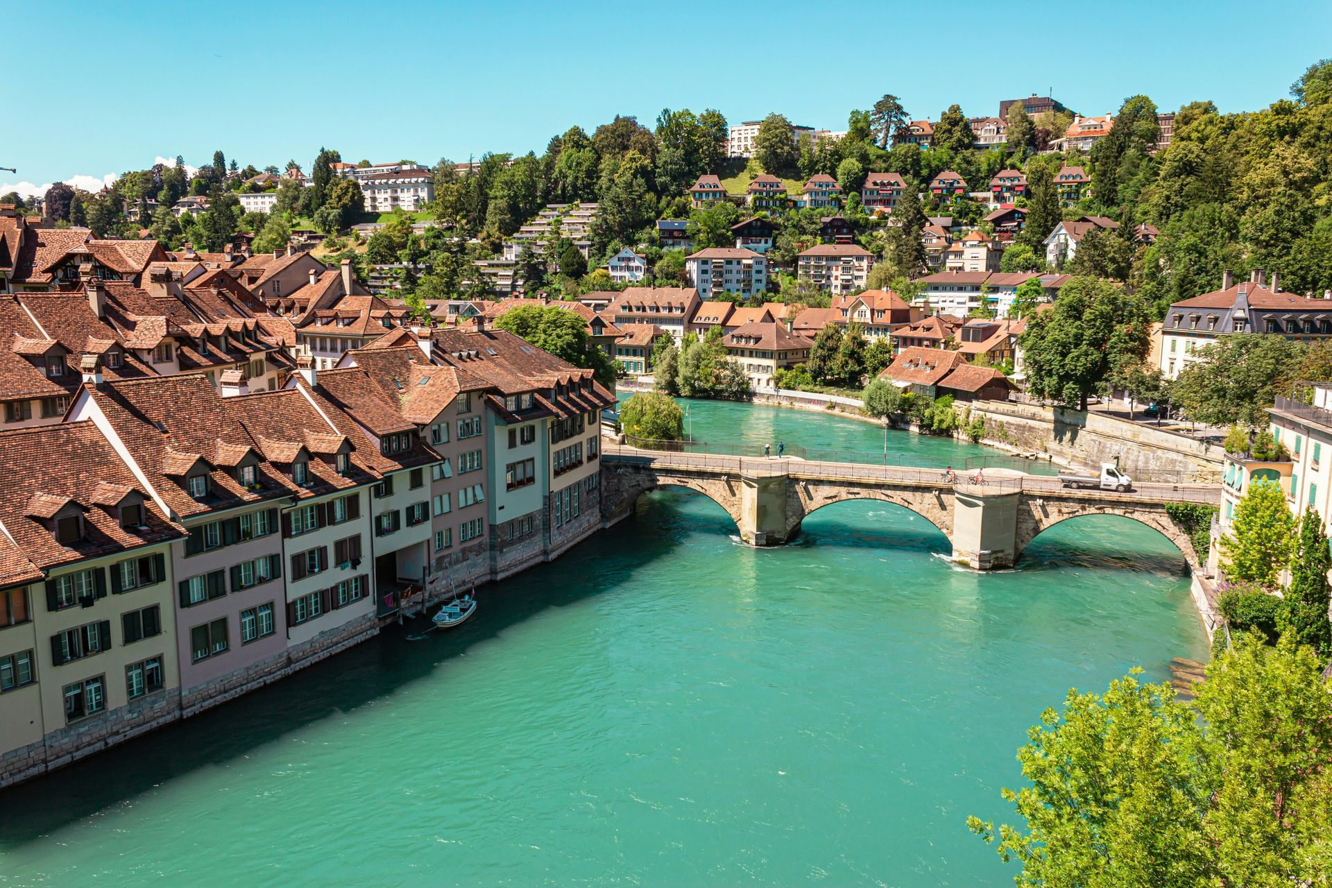 Bern, Switzerland: Buildings with red tile roofs along the turquoise Aare River, a stone bridge, and lush green hills.