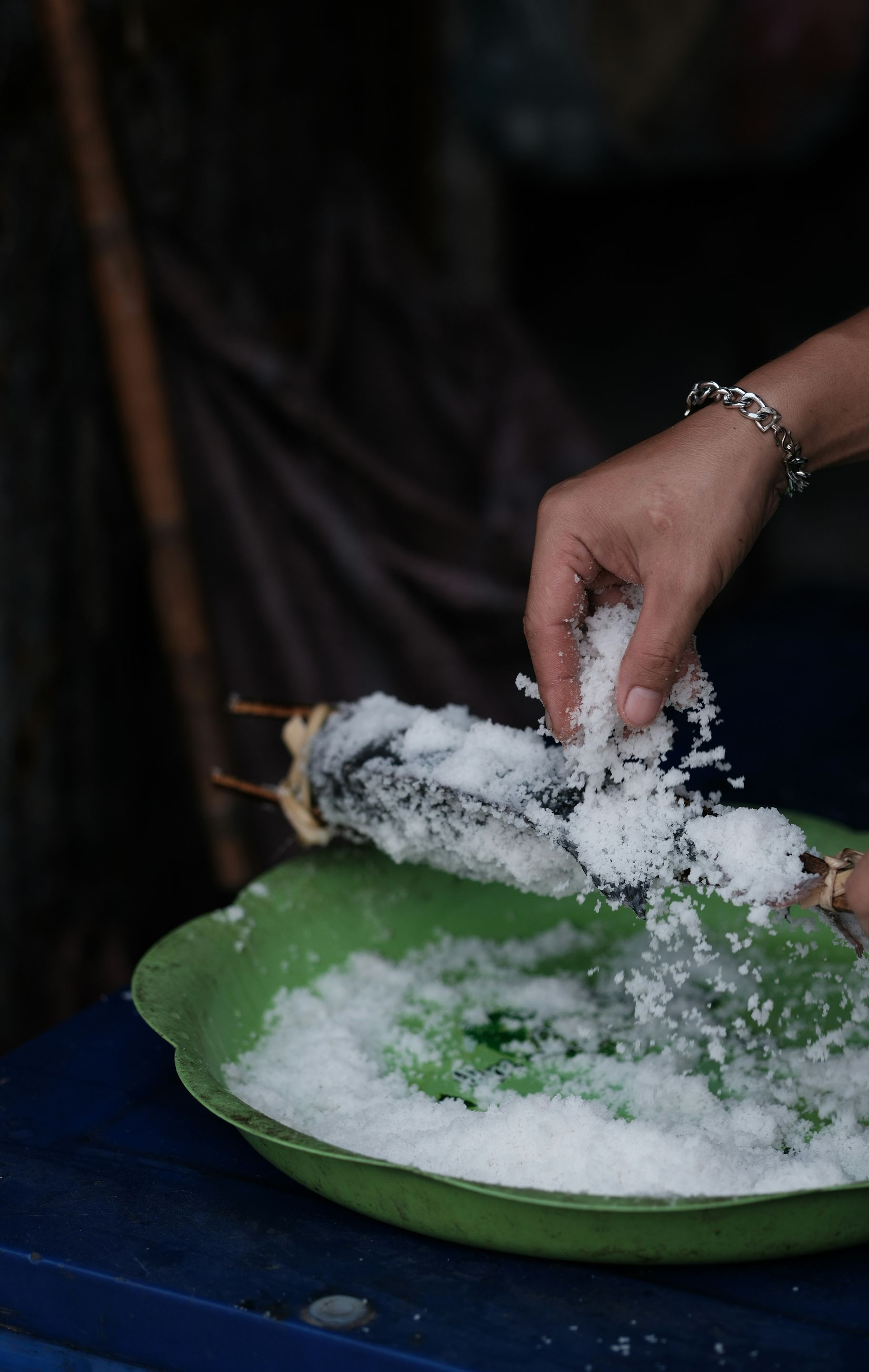 A person sprinkles salt onto fish held on a bamboo skewer over a green bowl in Laos.