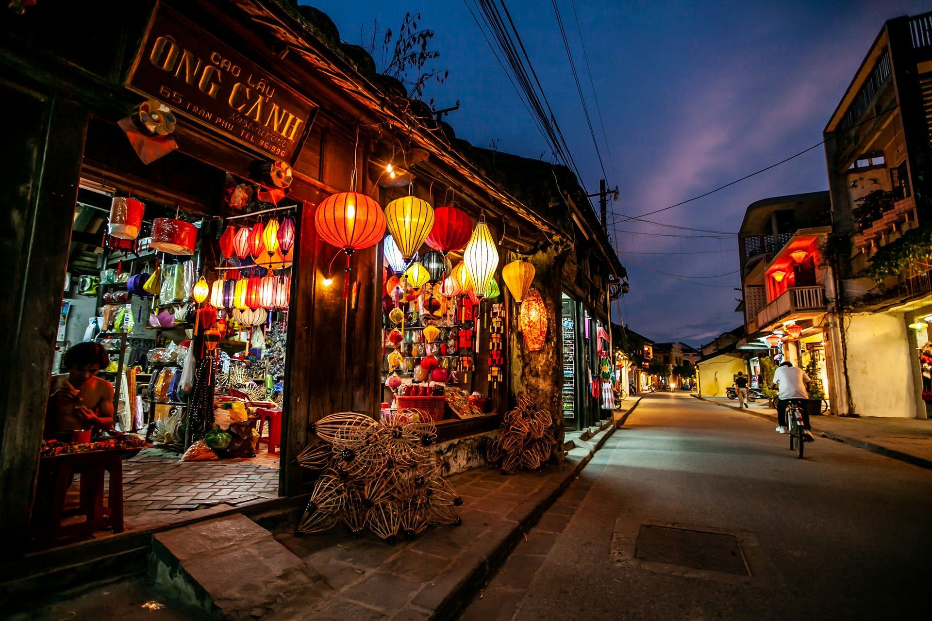 Street scene at dusk in Hoi An, Vietnam, with shops lit by colorful lanterns and a cyclist.