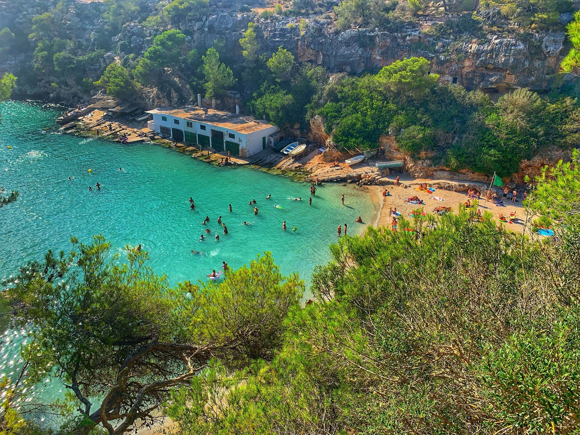 Turquoise bay with swimmers, sandy beach, white building, surrounded by green trees and rocky cliffs in Majorca, Spain.