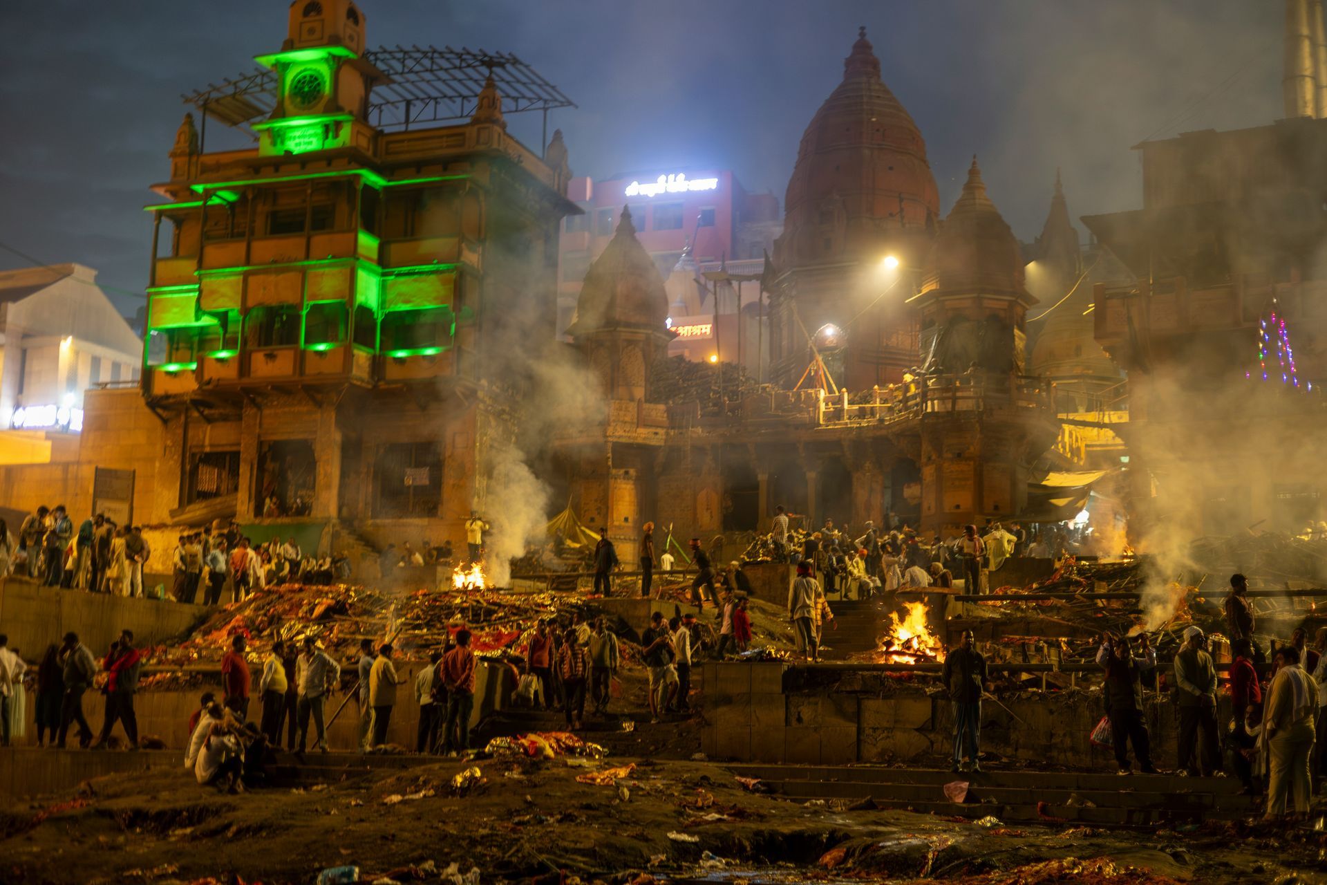 Cremation ghat in Varanasi, India, at night, with multiple funeral pyres burning, smoke and buildings in the background.
