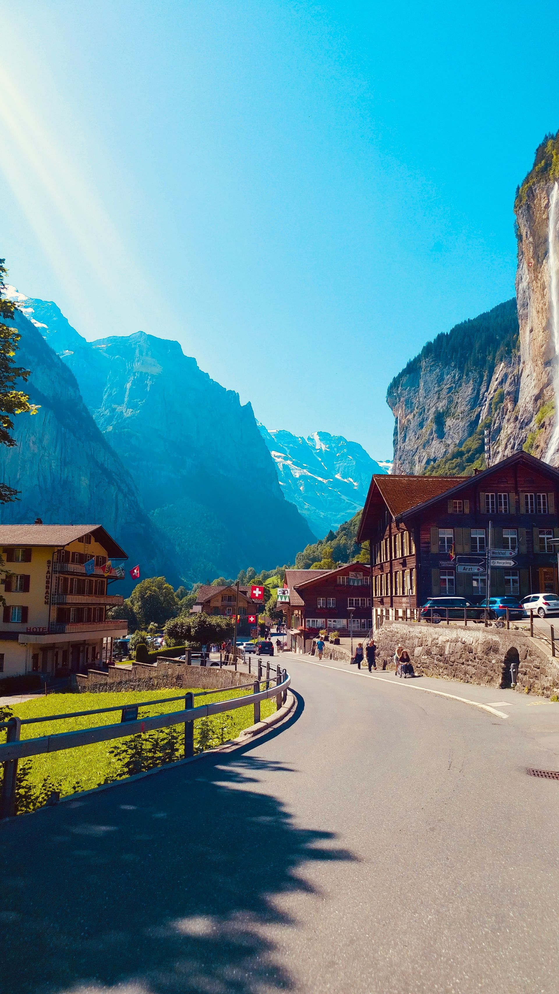 Mountain village scene with road, buildings, and waterfall under a bright blue sky in Lauterbrunnen.