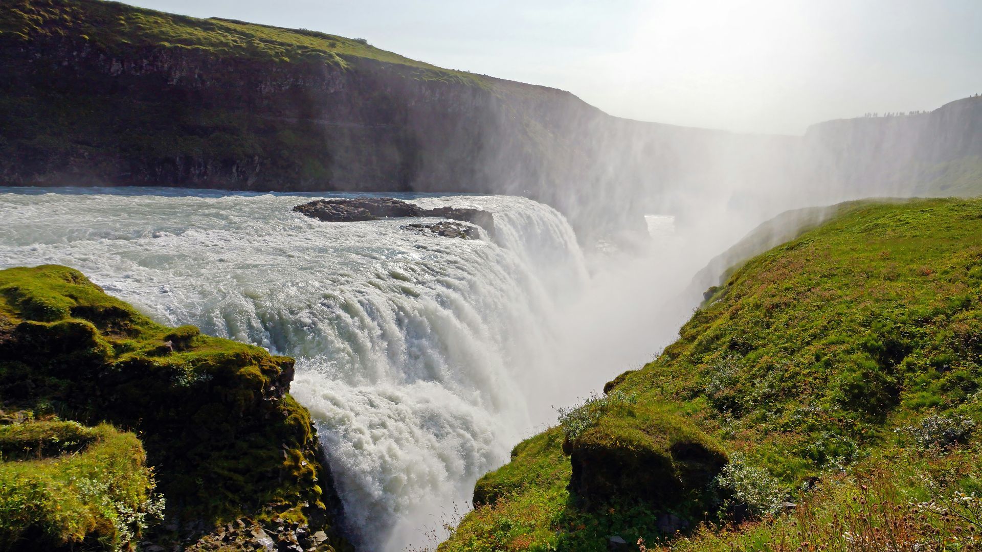 Gullfoss Falls cascading into a rocky gorge, with green banks under a hazy sky in Iceland.