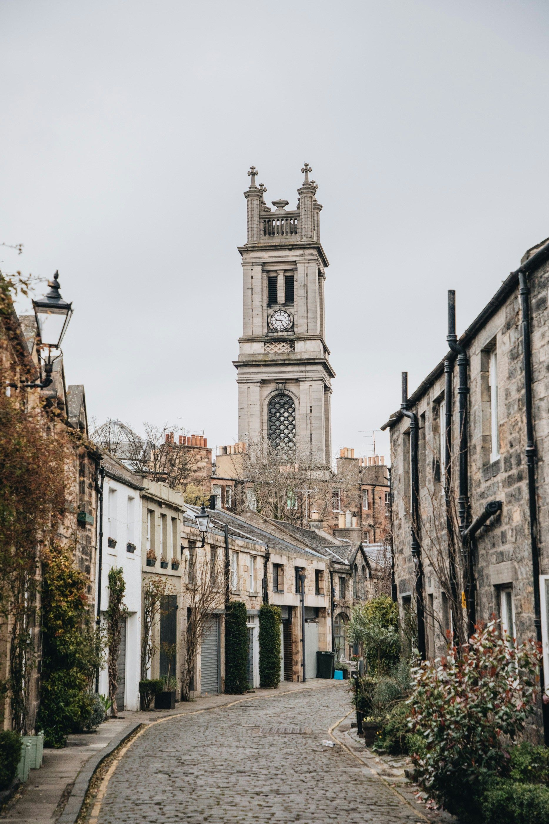 Cobblestone street in Edinburgh, Scotland, with stone buildings and a tall church tower on a cloudy day.