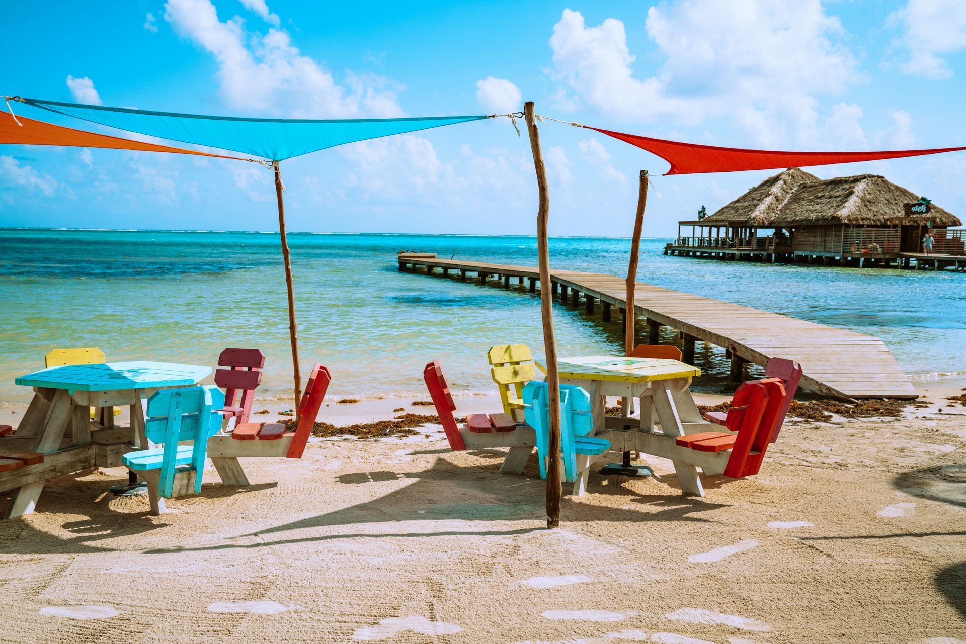 Colorful picnic tables with shade sails on a sandy beach. A wooden pier leads into the  water in Ambergris Caye, Belize.