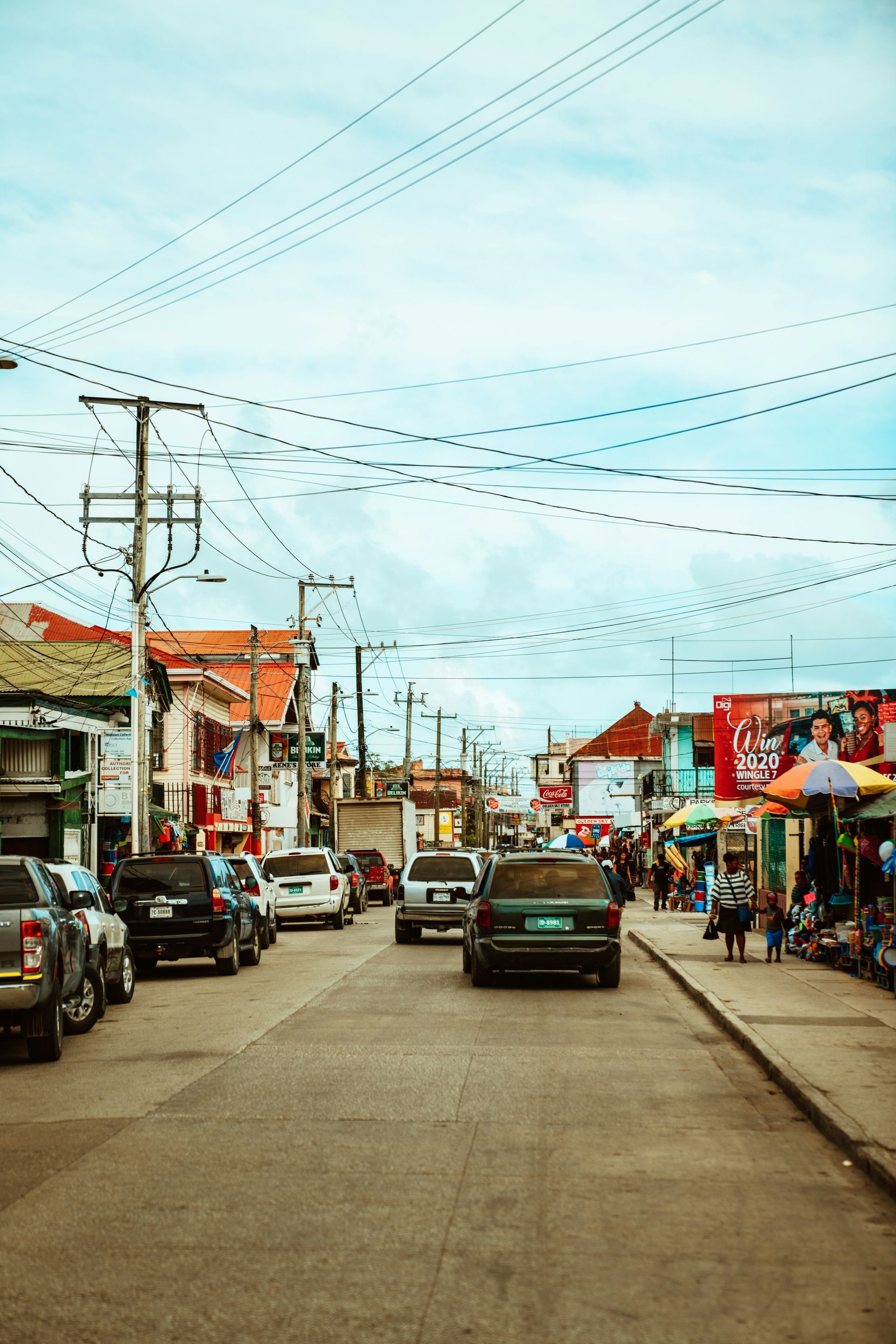 Street scene in a bustling town with cars and buildings, and people walking along the sidewalks in Belize City.