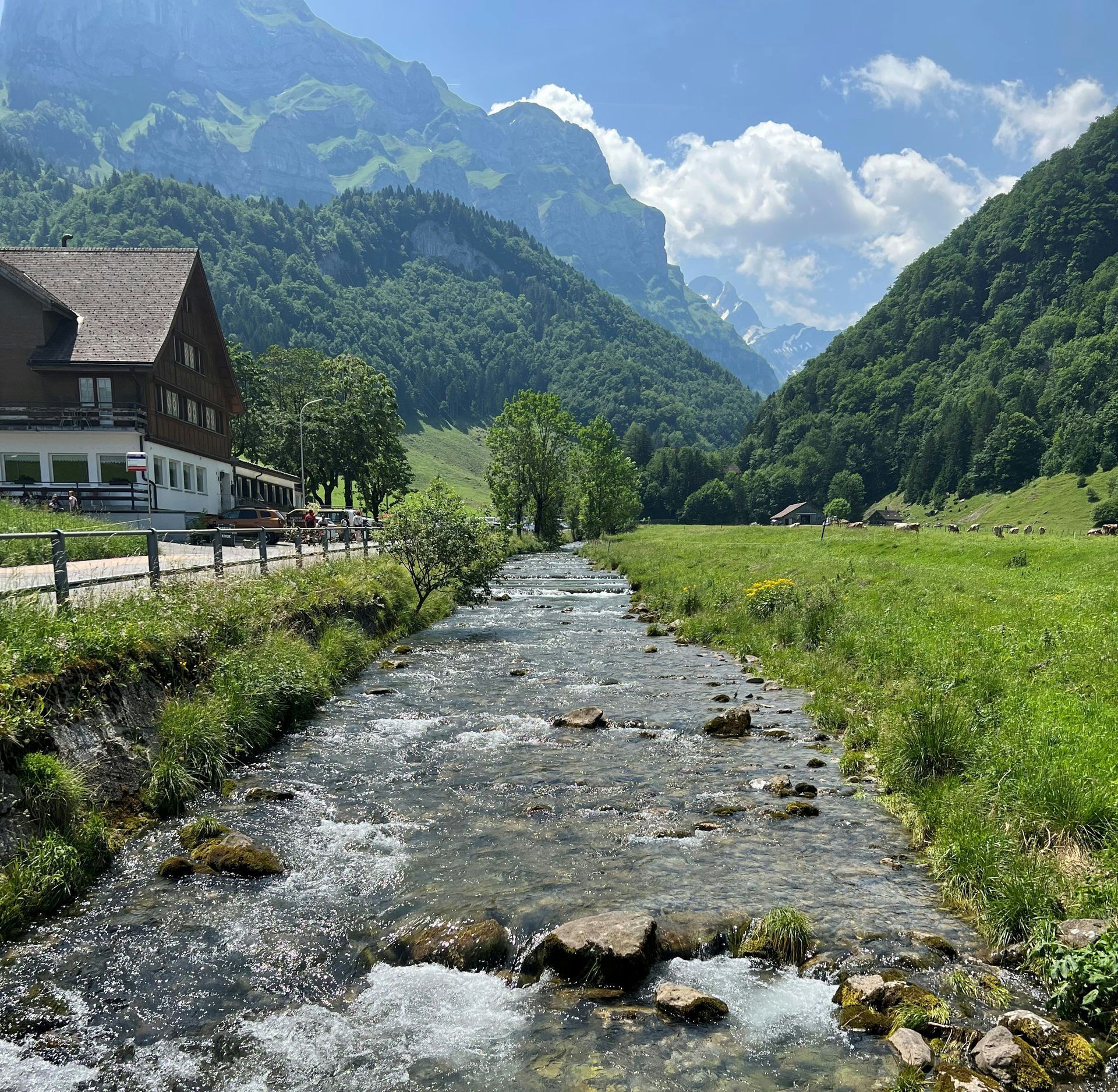 A river flows through a green valley, with a mountain backdrop and a chalet on the left in Schwende, Switzerland.