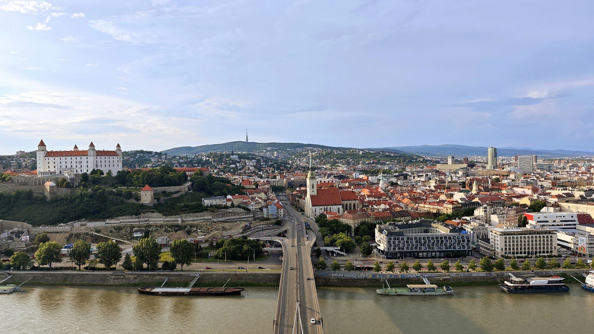 Bratislava cityscape with castle, bridge, and Danube River under a cloudy sky.