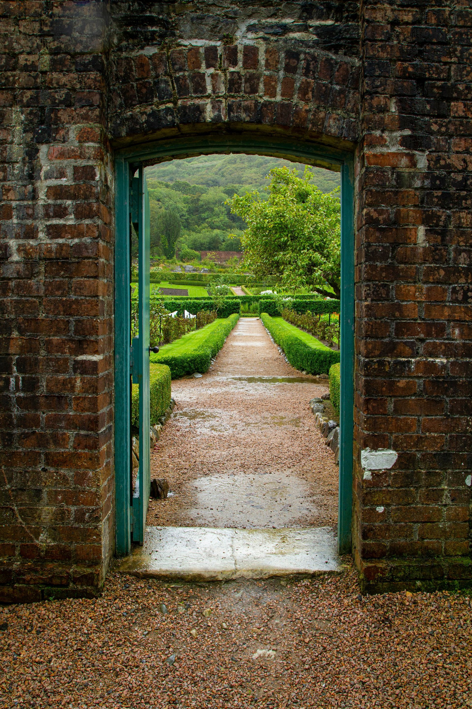 Brick archway frames a garden path, lined with hedges, leading to lush greenery in Connemara, Ireland.