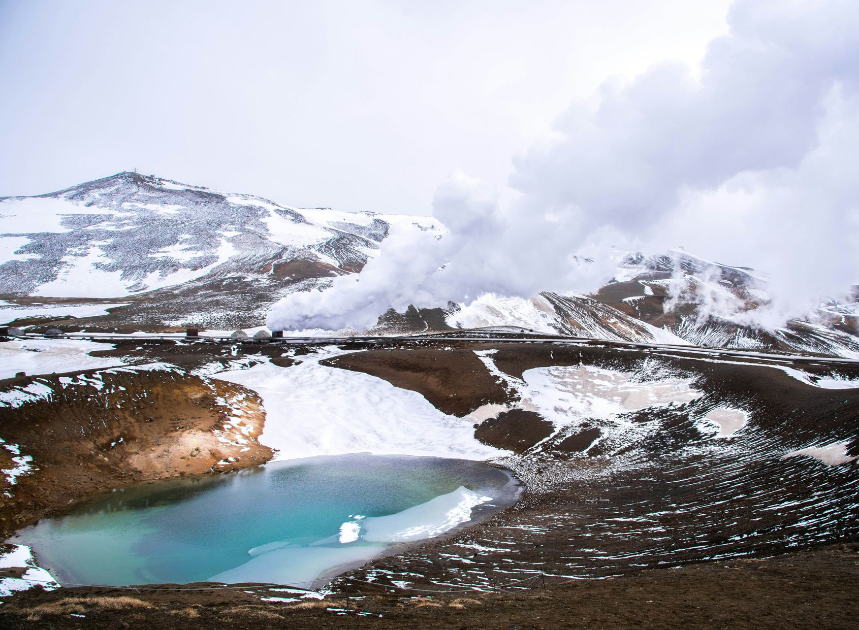 Snowy landscape with a turquoise Askja lake and steam rising from geothermal vents in Iceland.