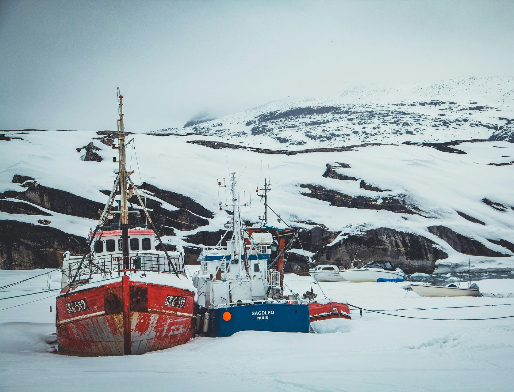 Red and blue boats docked on snowy shore; snow-covered hills in the background in Disko Bay, Greenland.
