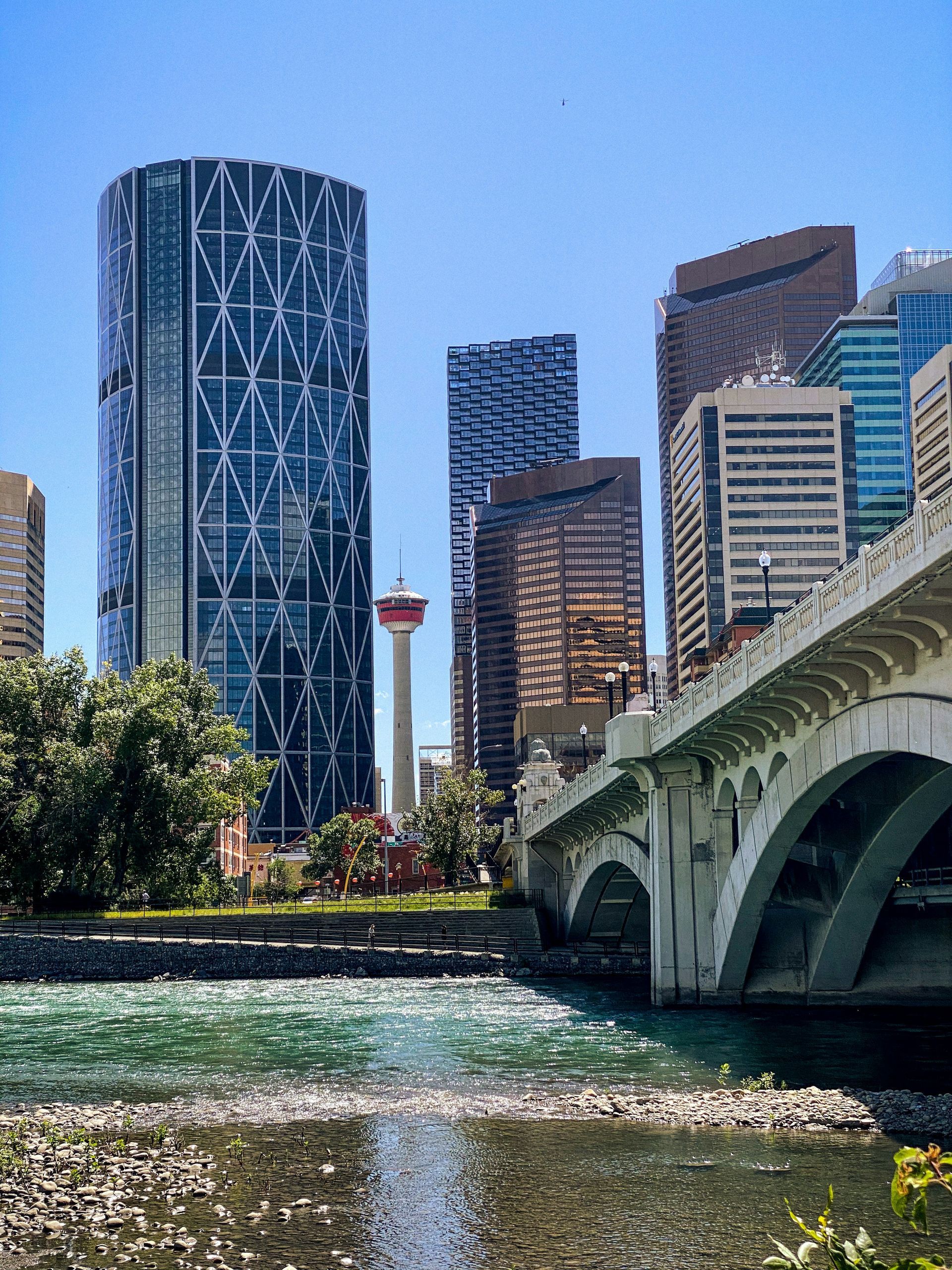 Downtown Calgary skyline with skyscrapers, the Calgary Tower, and a bridge arching over the Bow River under a clear blue sky.