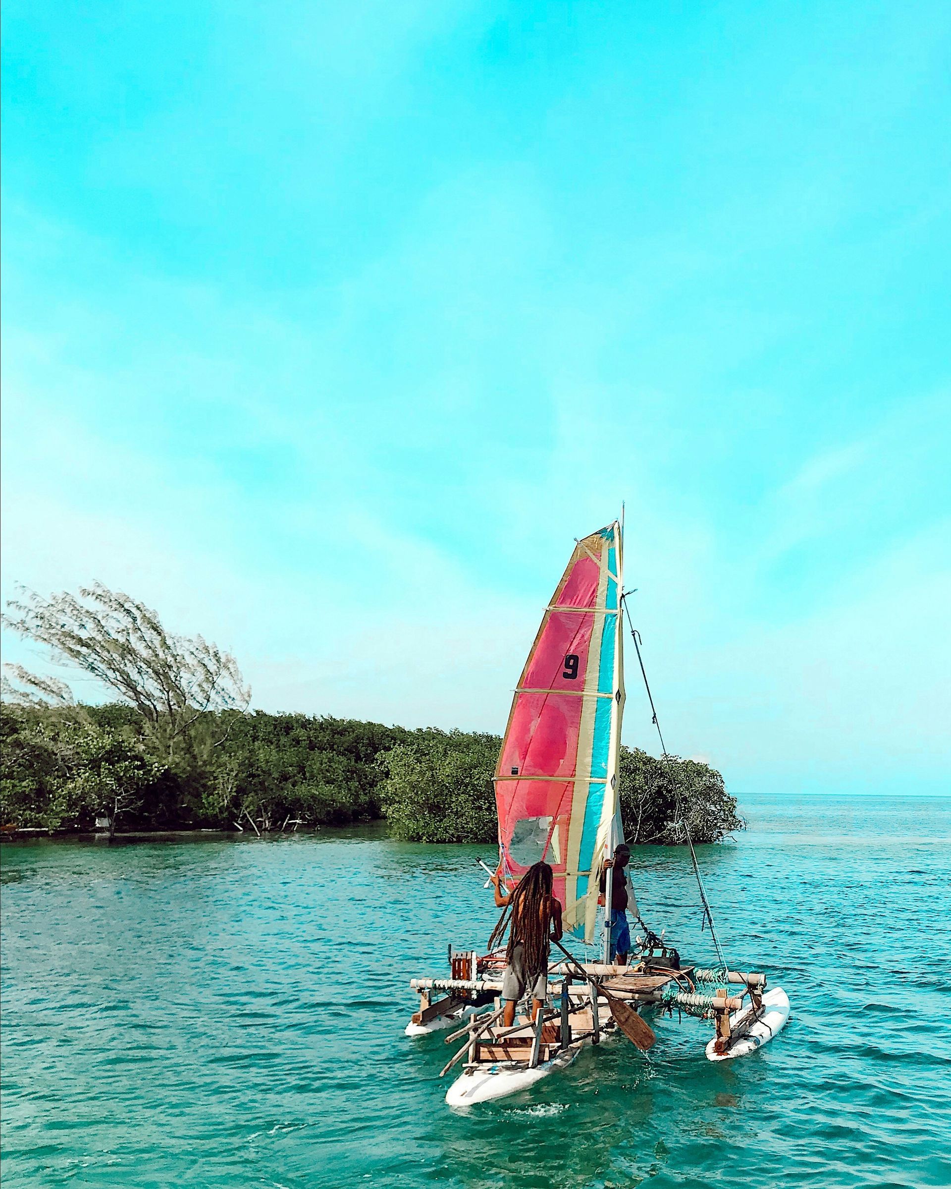 A catamaran sailboat with a colorful sail glides across turquoise water in Caye Caulker, Belize.