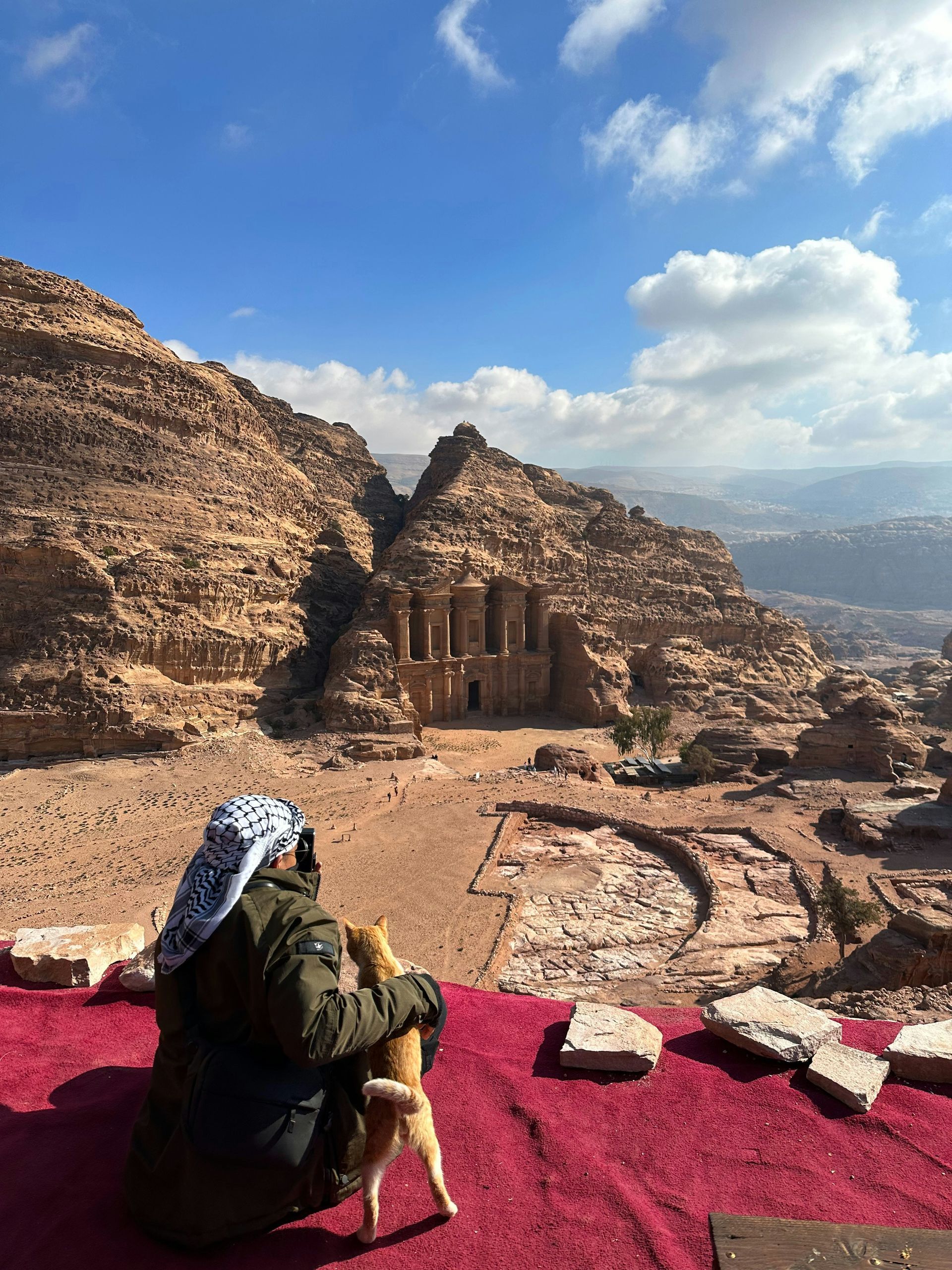 Person in traditional head covering sits with a dog, gazing at Petra's Monastery, carved into sandstone cliffs.