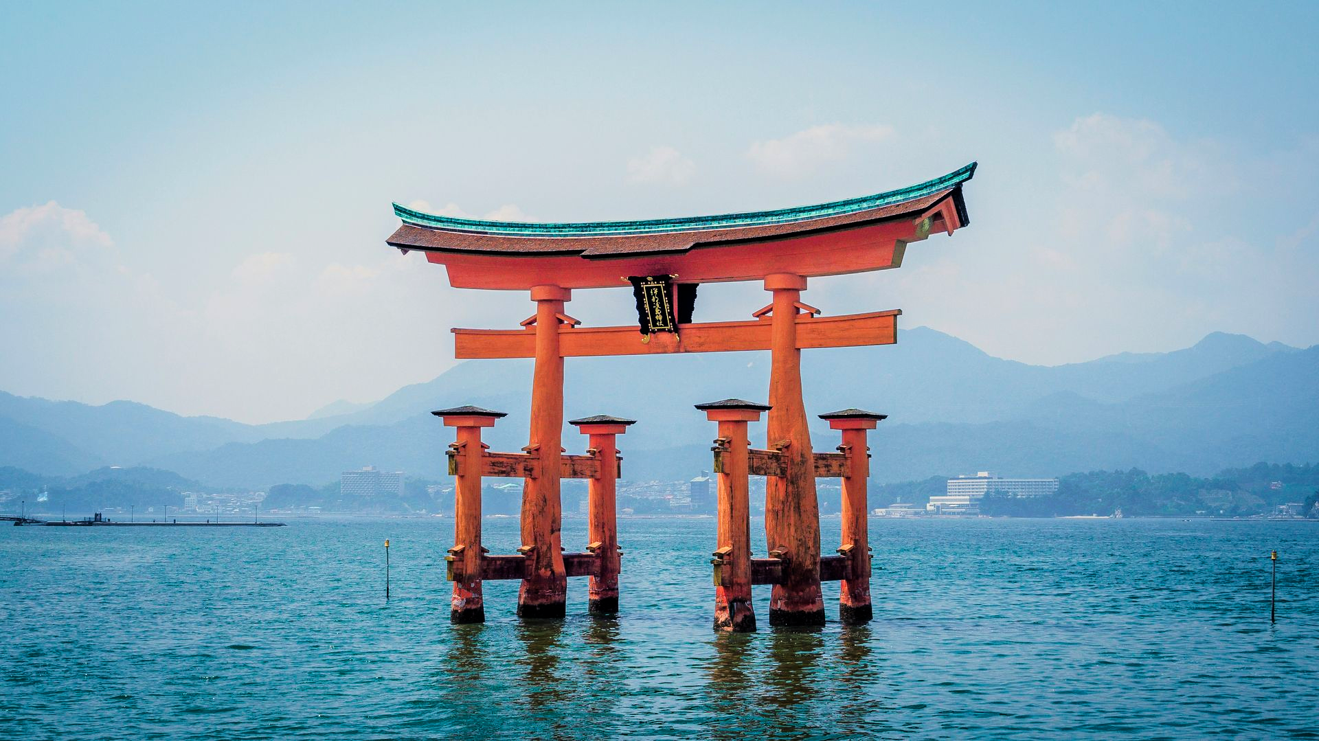 Torii gate in water, orange with green roof, mountains in the distance in Hiroshima, Japan.