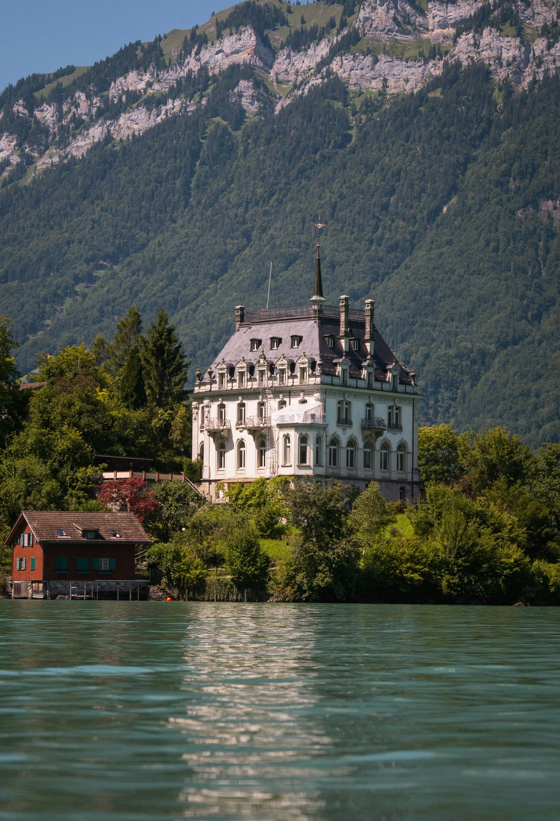 The white Seeburg Castle on Lake Brienz, with a red house to its left. Mountains in the background in Switzerland.