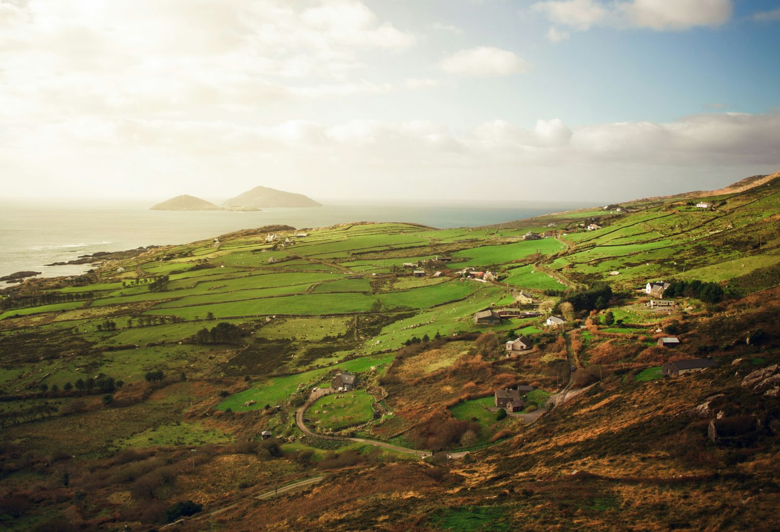 Rolling green hills meet the ocean; small houses dot the landscape under a bright sky in Ireland.