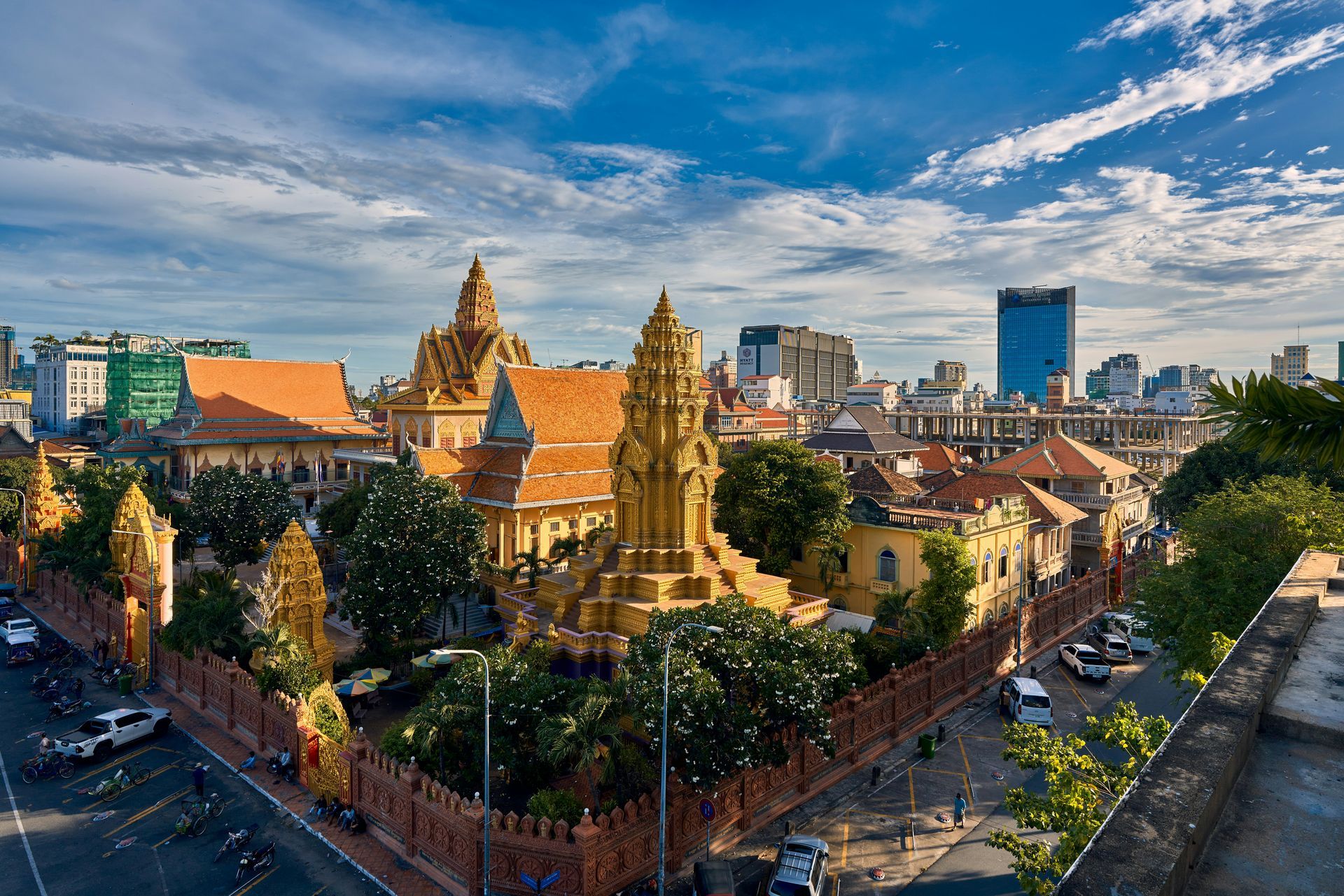 View of Phnom Penh, Cambodia, with gold-roofed buildings, ornate walls, and a cityscape under a partly cloudy sky.