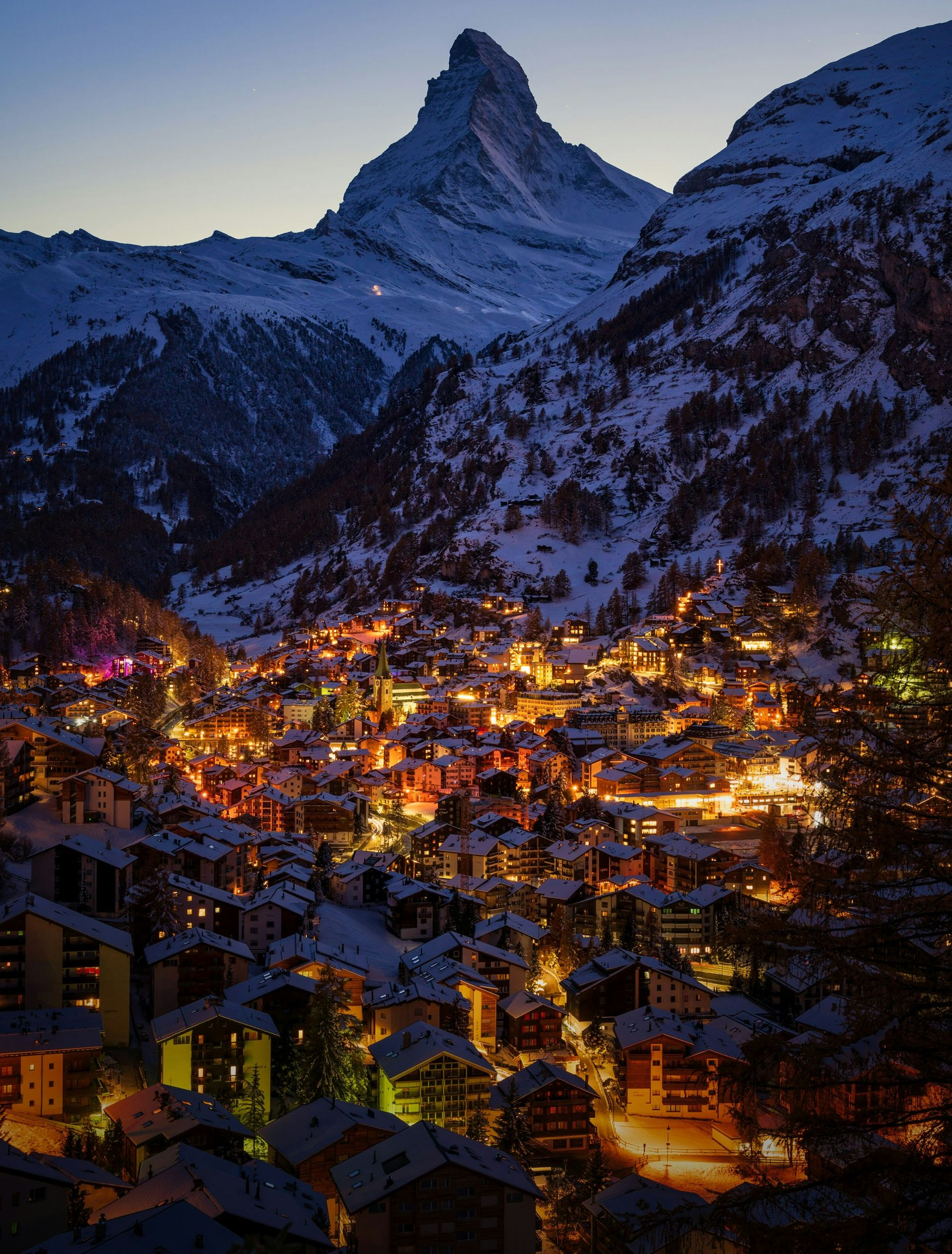 Snow-covered Swiss village at dusk, lit by warm lights, with Matterhorn mountain in the background in Zermatt, Switzerland.