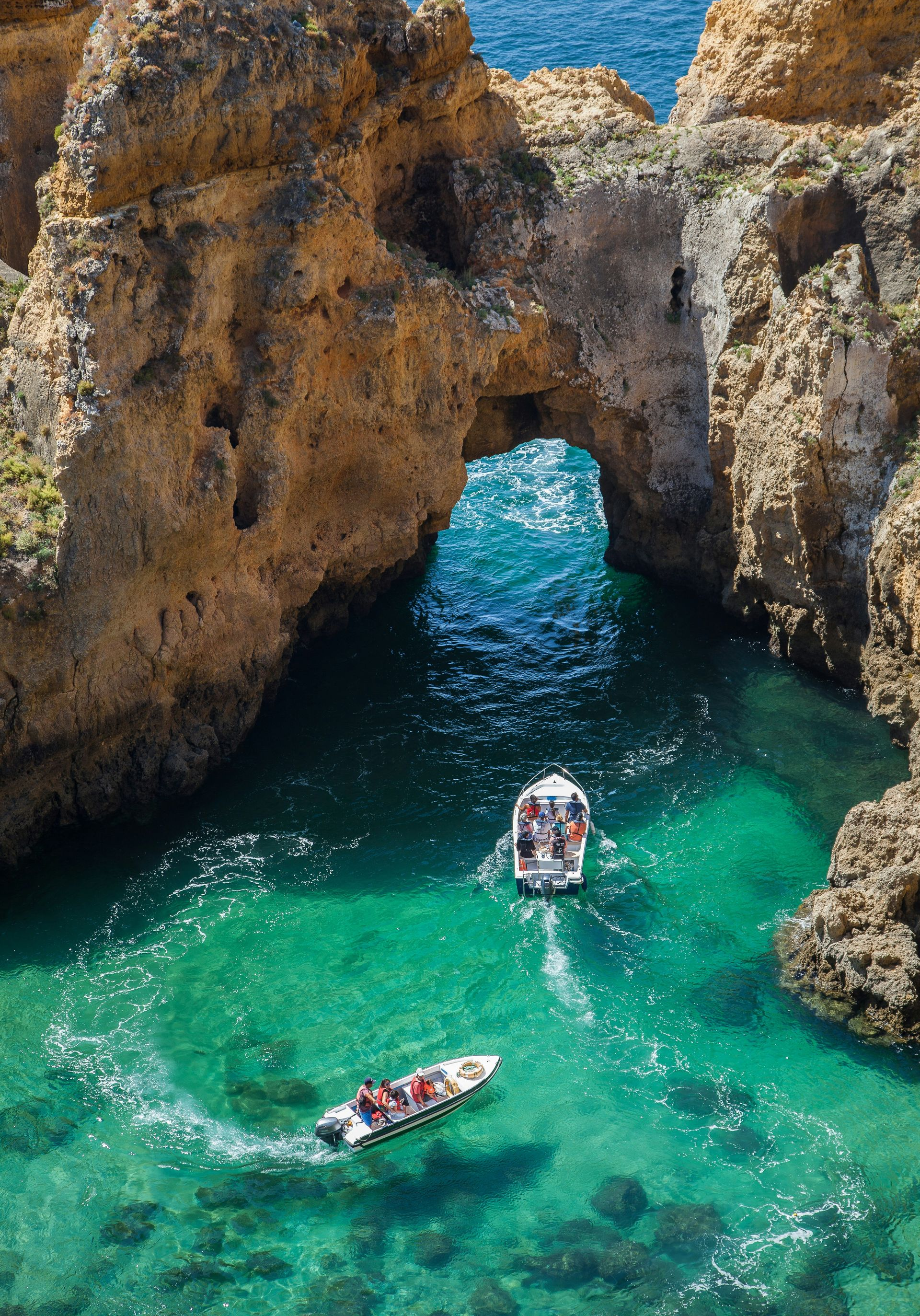 Two boats navigate a turquoise sea through a natural archway in a rocky coastline in The Algarve, Portugal.