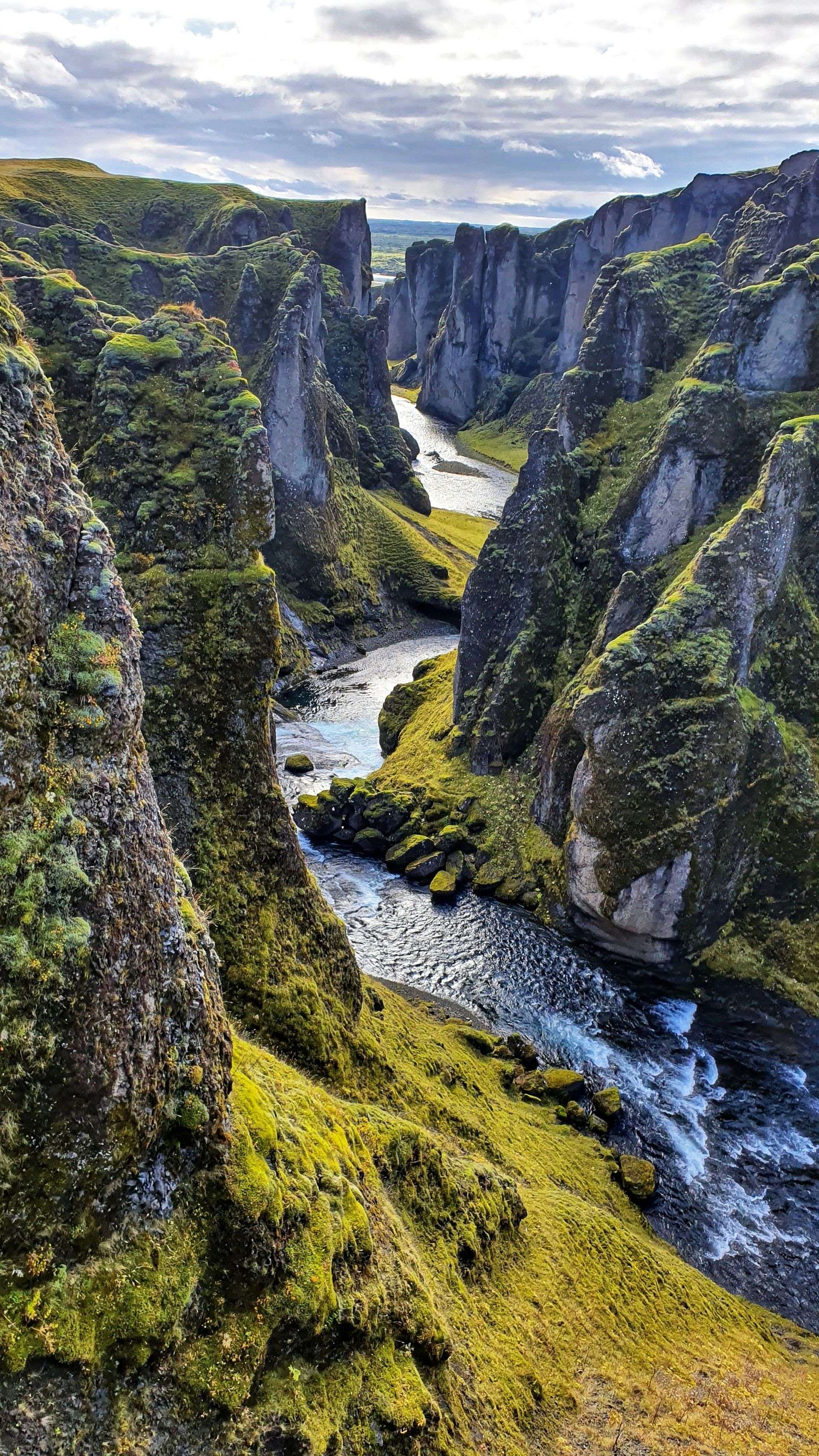 A river winds through a mossy canyon with rocky walls, under a cloudy sky at Fjaðrárgljúfur, Iceland.