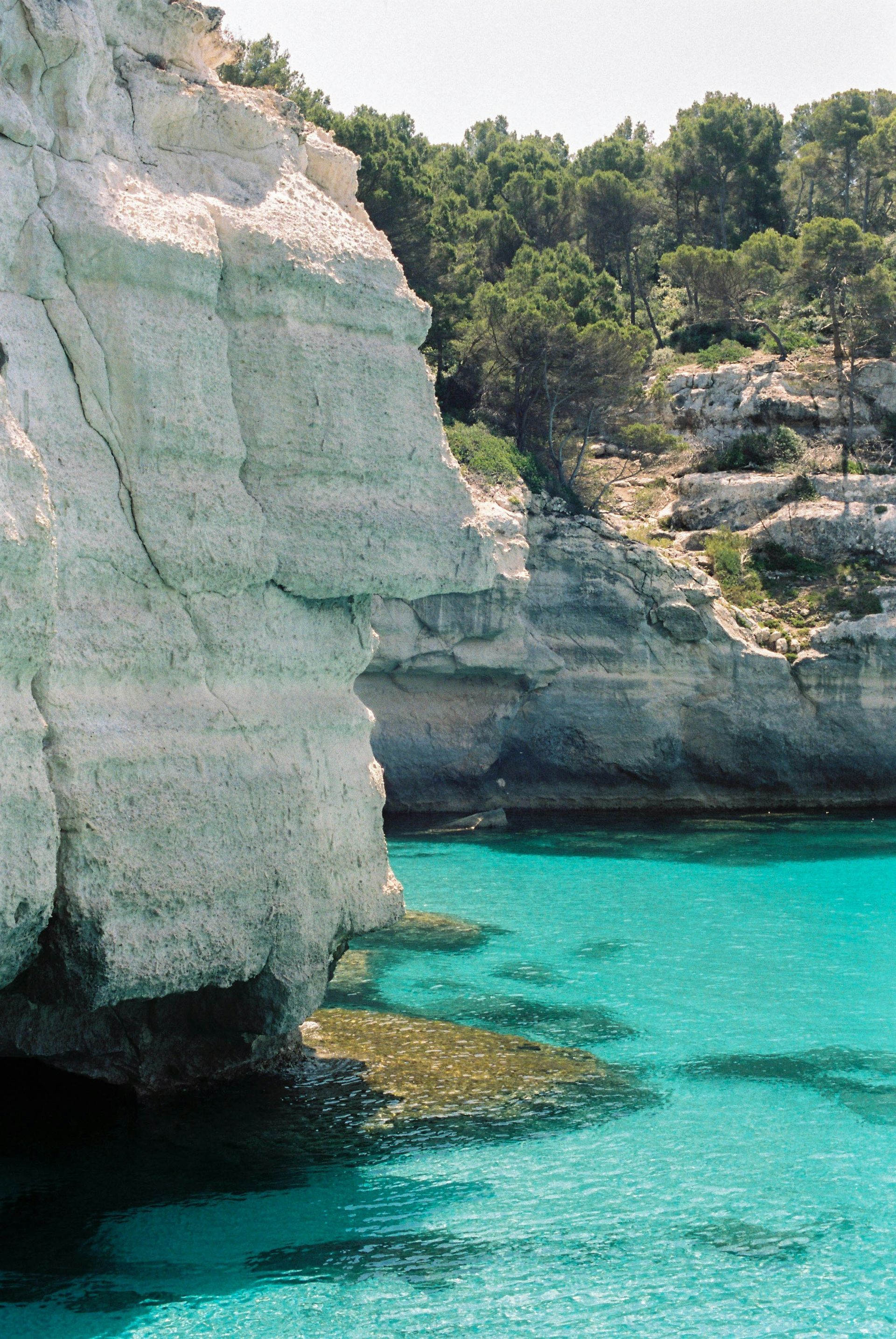 A cliff overlooking a body of water with trees in the background in Menorca, Spain.
