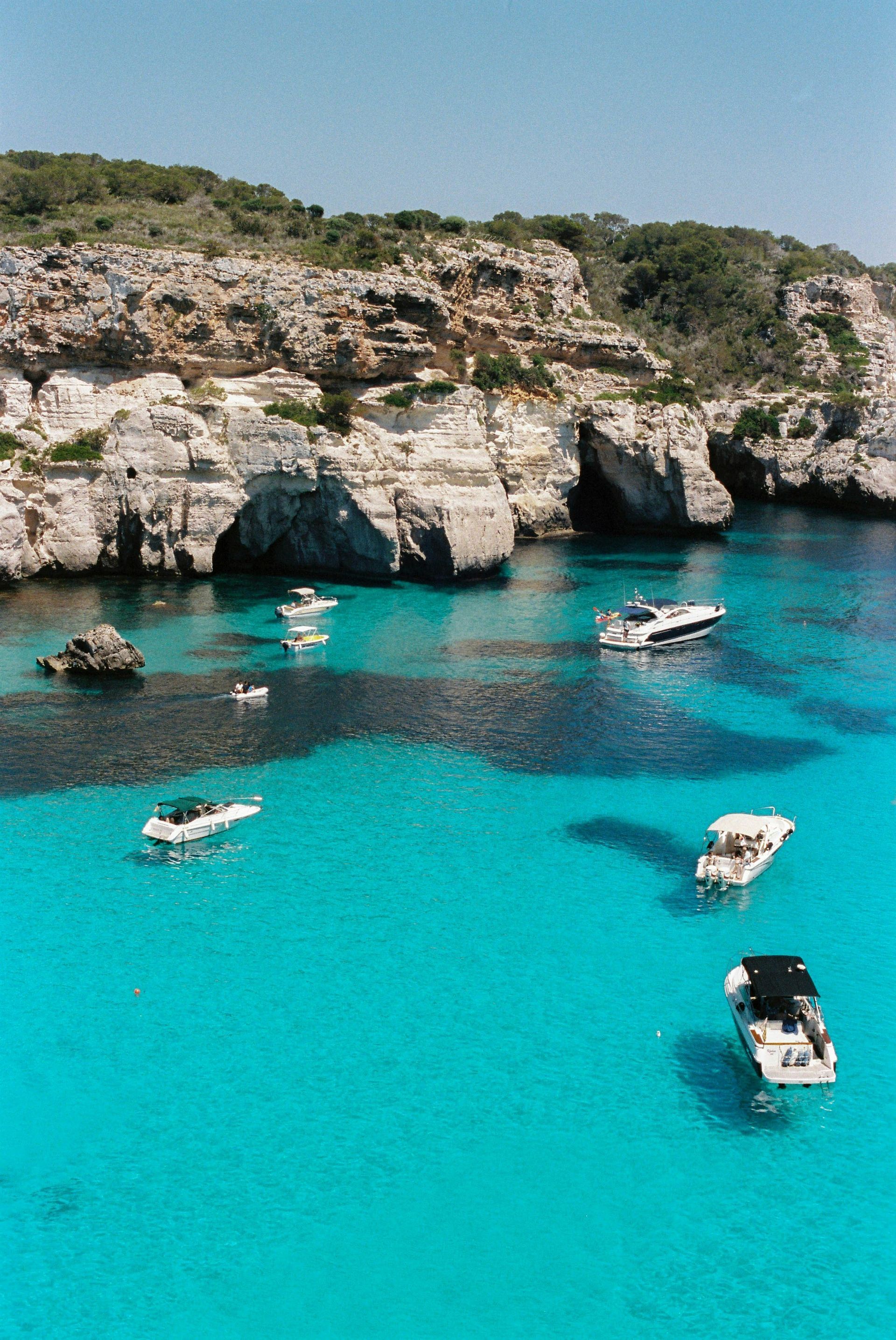 Boats float in bright turquoise water near a rocky coastline on a sunny day in Menorca, Spain.