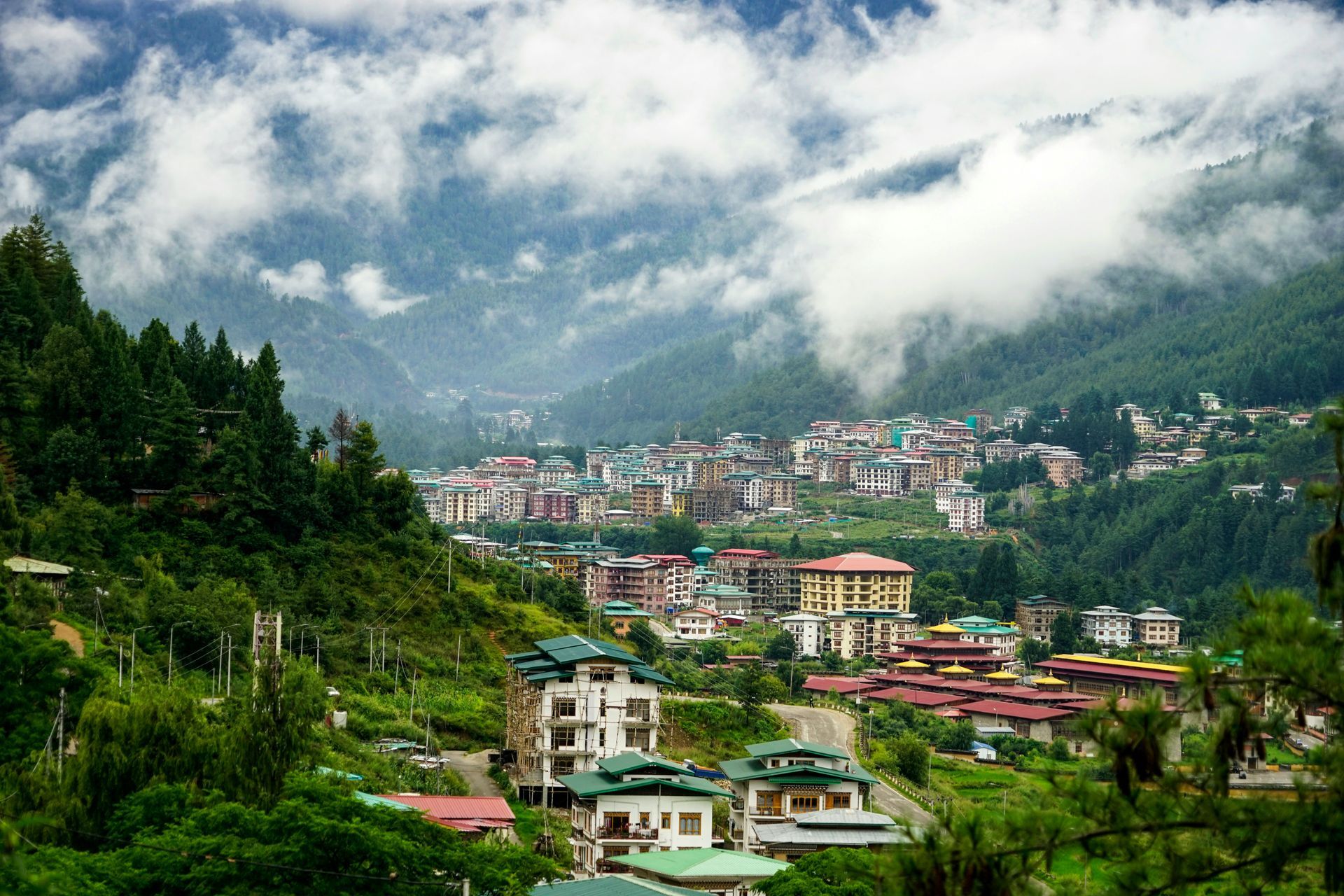 Mountain town of Thimphu in Bhutan, with buildings nestled in a valley, shrouded in clouds. Green trees cover the slopes.